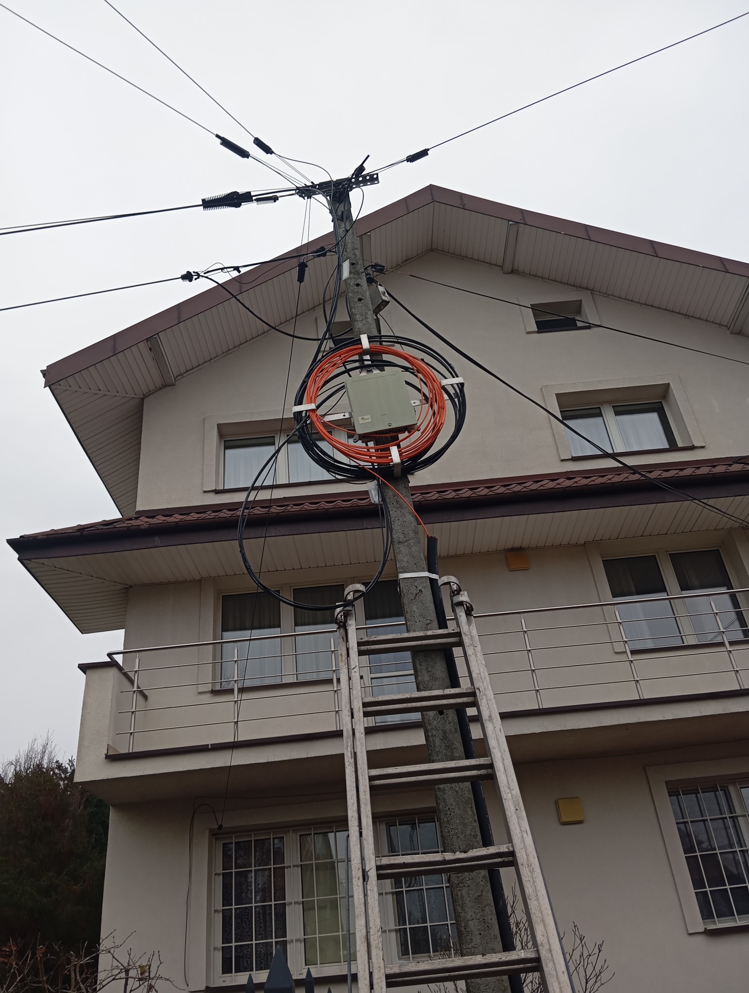Słup energetyczny z zamontowaną skrzynką i zwiniętymi kablami, drabina oparta o słup, na tle dwupiętrowego budynku z balkonem.