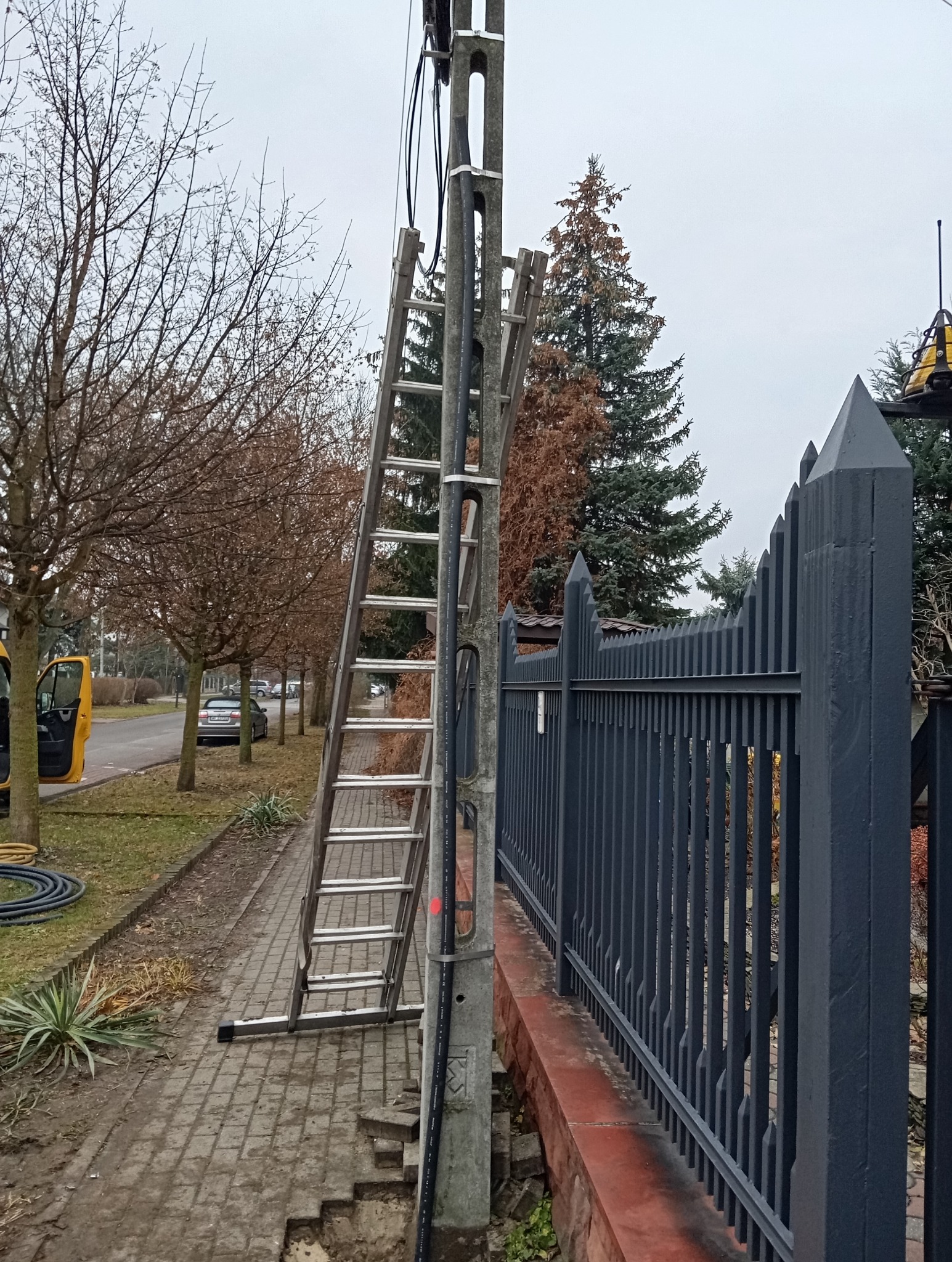 Aluminiowa drabina oparta o betonowy słup energetyczny na chodniku z kostki, obok wykop pod kable, w tle czarny metalowy płot i drzewa.