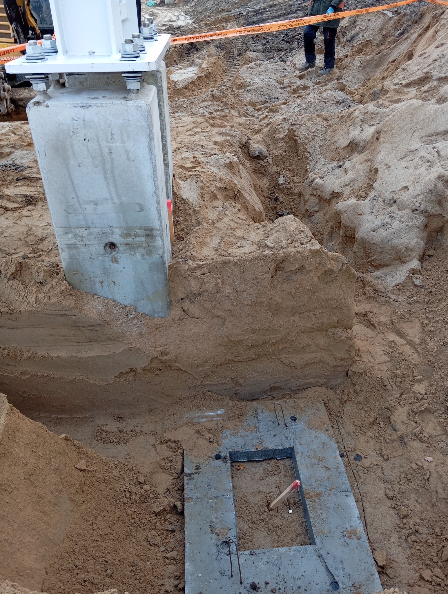 Betonowy słup mocowany na fundamencie w wykopie, widoczne ślady po pracy koparki i taśma ostrzegawcza nad wykopem.