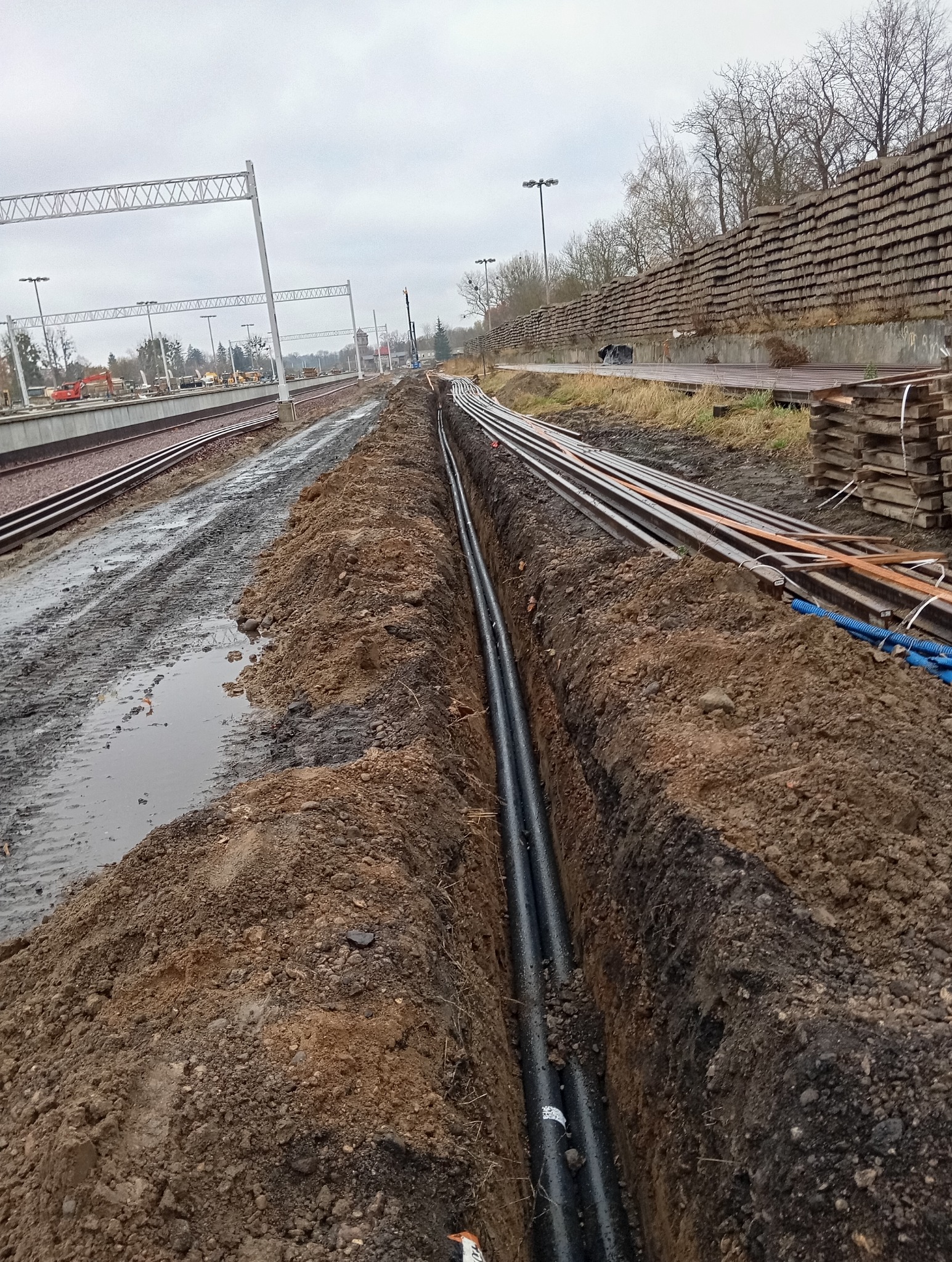 Wykopany rów z ułożonymi czarnymi rurami wzdłuż torów kolejowych, widoczne błoto i urządzenia budowlane w tle.