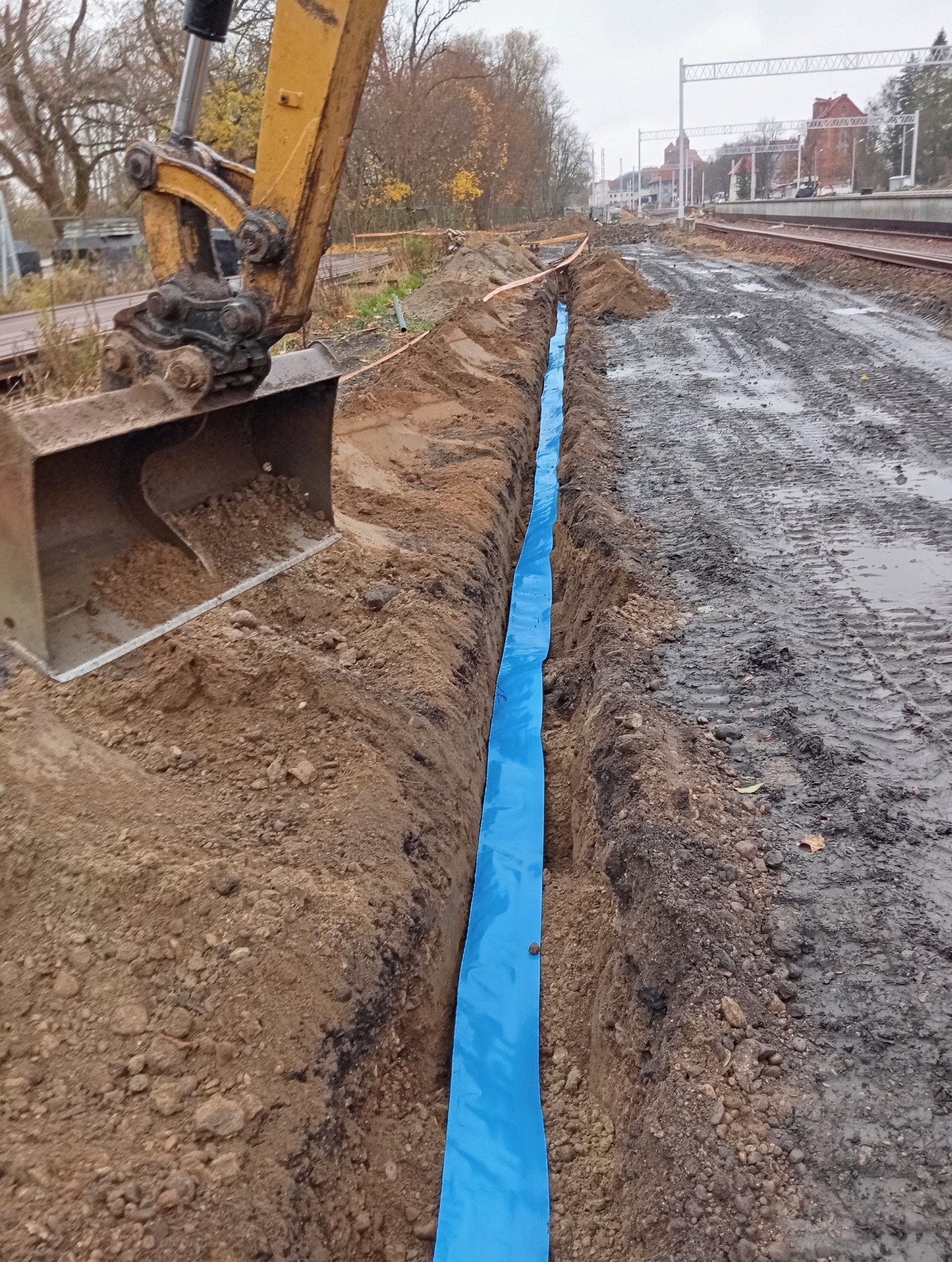 Wykopany wąski rów z niebieską folią ochronną w ziemi, obok torów kolejowych, z koparką w tle.
