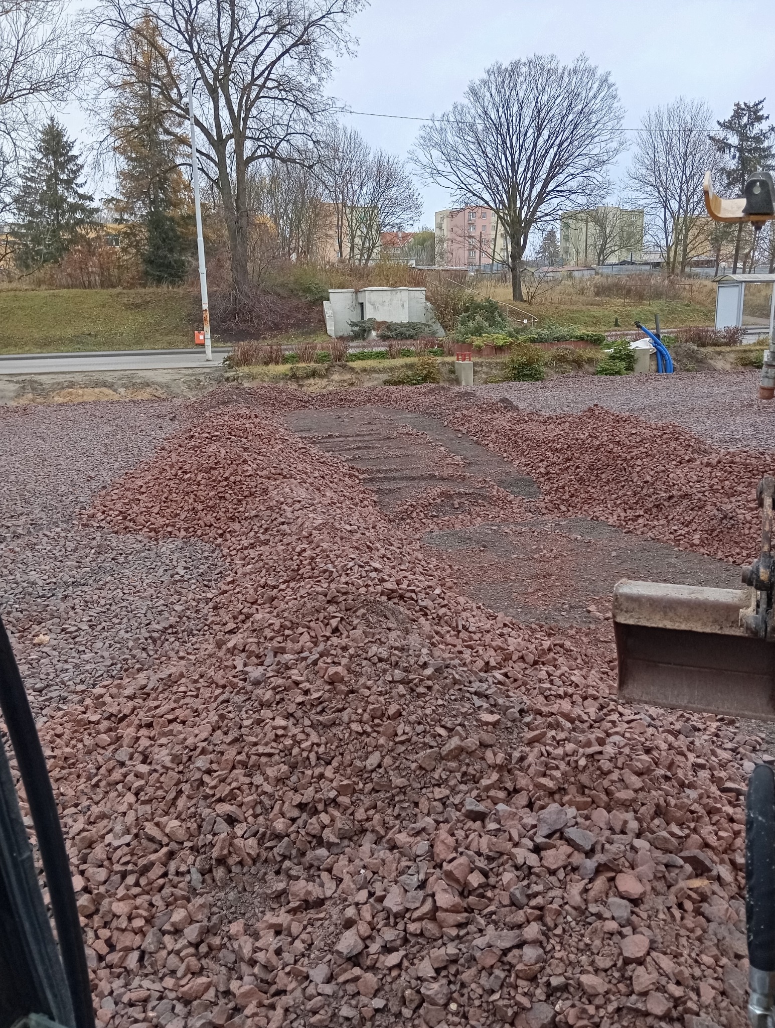Rozplantowany teren z czerwonym gruzem, widoczny fragment łyżki koparki, w tle zabudowa miejska i drzewa bez liści.