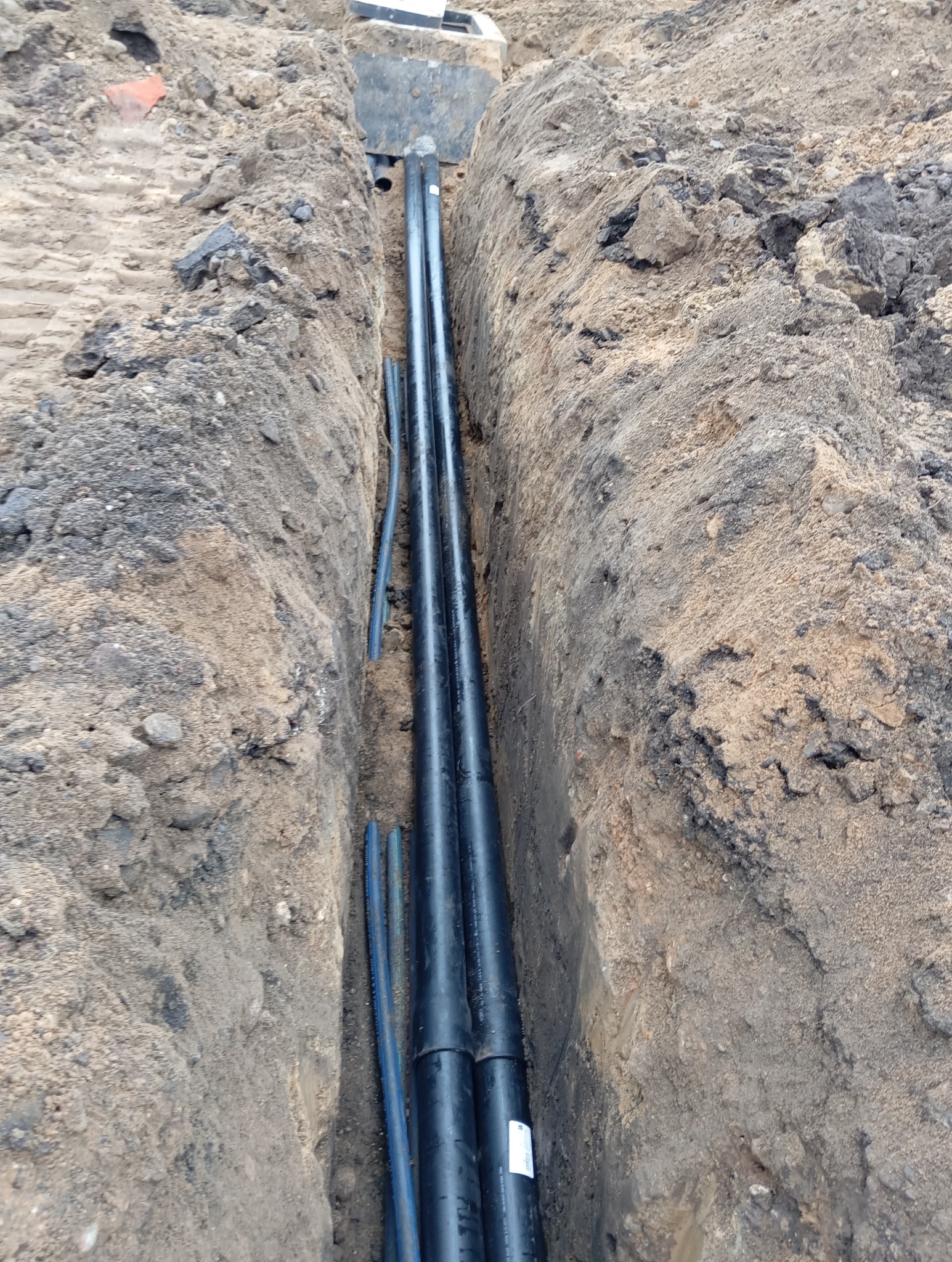 Wykopany rów z ułożonymi rurami instalacyjnymi, widoczne ściany wykopu z ziemi i piasku.