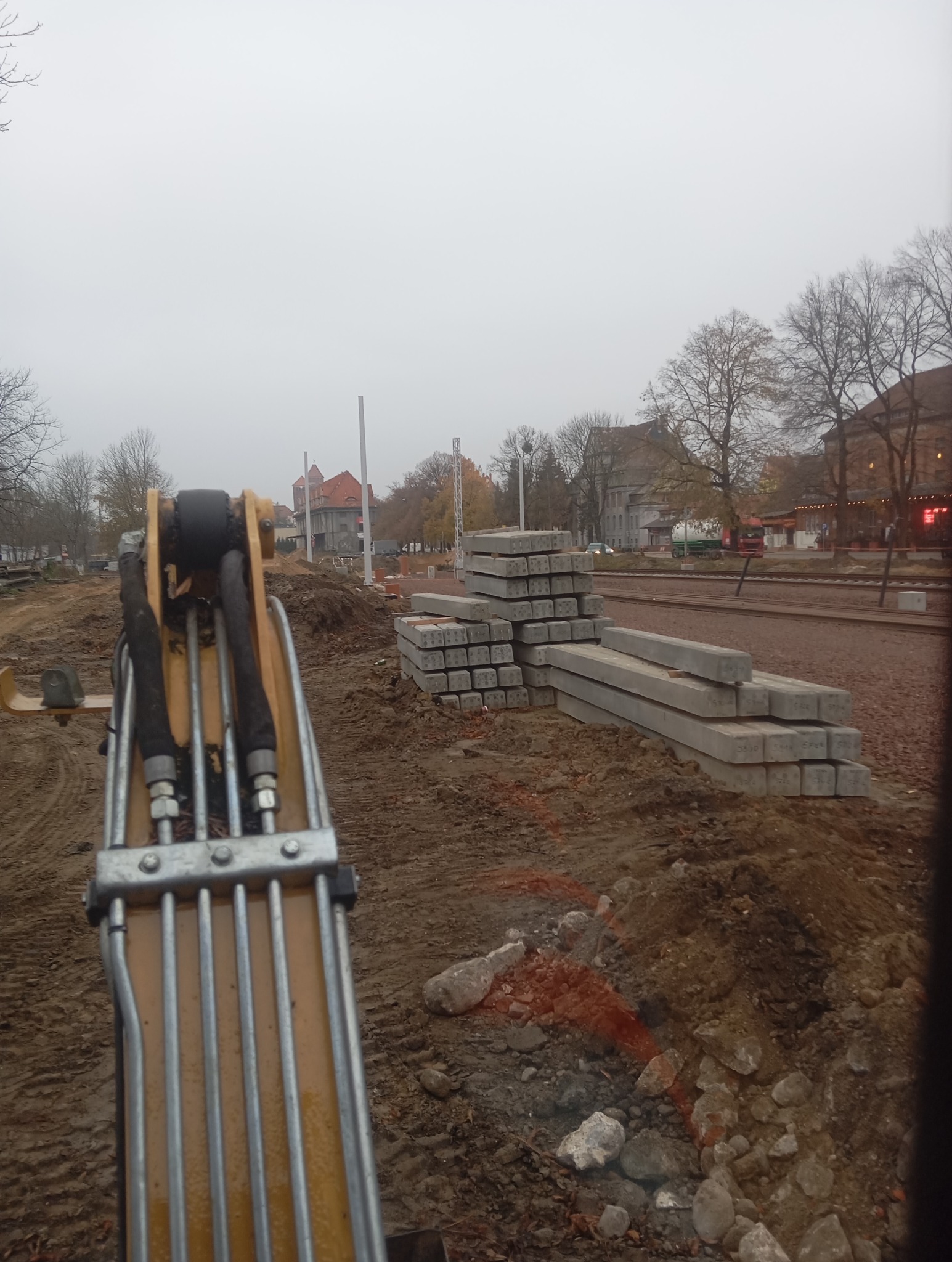 Ramię koparki z widocznymi przewodami hydraulicznymi na tle wykopu, ułożonych betonowych elementów i budynków w oddali, pochmurny dzień.