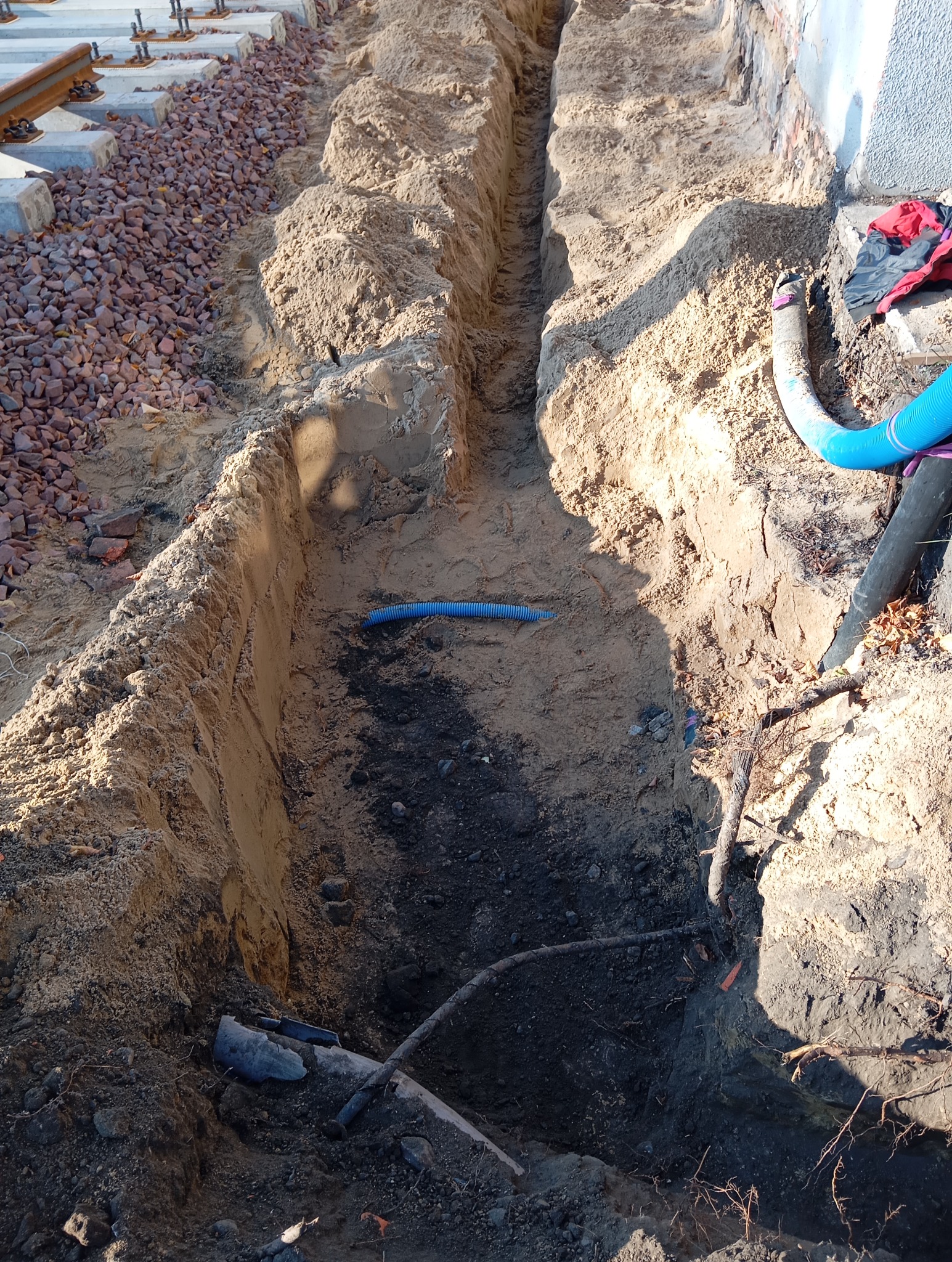 Długi, wąski wykop w piaszczystej ziemi, z widocznymi niebieskimi rurami karbowanymi wewnątrz, obok fundamentów budynku i podsypki z kamieni. Wykop częściowo wypełniony ziemią.