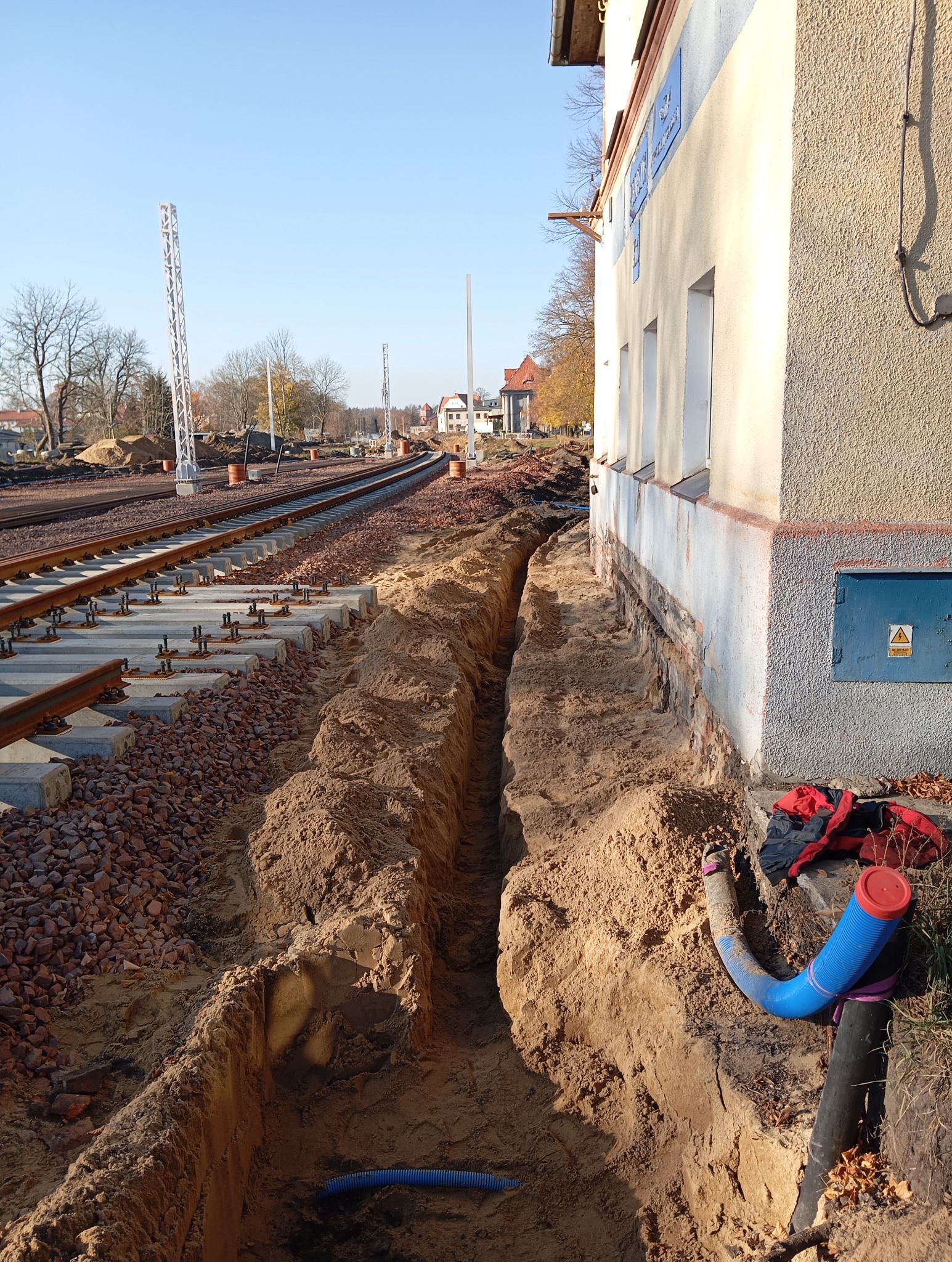 Wkop pod instalacje wzdłuż budynku dworca kolejowego, widoczne rury instalacyjne i fragment torów.