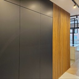 Krzysztof Pęczek, F.H.U. AGMA - Nowoczesna szafa w zabudowie z matowymi, grafitowymi frontami i dekoracyjnymi lamelami z drewna. W tle widać fragment kuchni i okno.