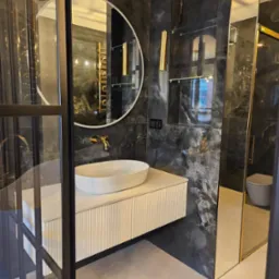Nowoczesna łazienka z umywalką nablatową, okrągłym lustrem i czarnymi płytkami imitującymi marmur. Widoczna kabina prysznicowa ze złotymi detalami.
