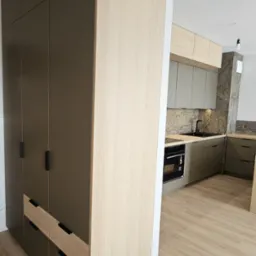 Nowoczesna kuchnia z zabudową: wysoka szafa w kolorze taupe z drewnianymi akcentami i jasne szafki kuchenne z kamiennym backsplashiem. Minimalistyczny design.