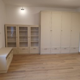 Krzysztof Pęczek, F.H.U. AGMA - Jasna zabudowa meblowa: szafa, witryna i komoda z szufladami, tworząca spójną aranżację wnętrza na drewnianej podłodze.