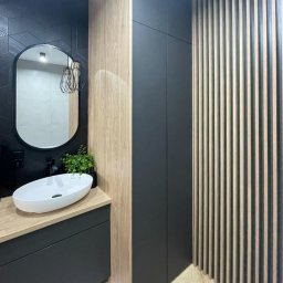 Krzysztof Pęczek, F.H.U. AGMA - Nowoczesna łazienka z owalnym lustrem, umywalką nablatową i drewnianymi lamelami. Ciemne płytki i szafki tworzą elegancki kontrast z jasnym drewnem.