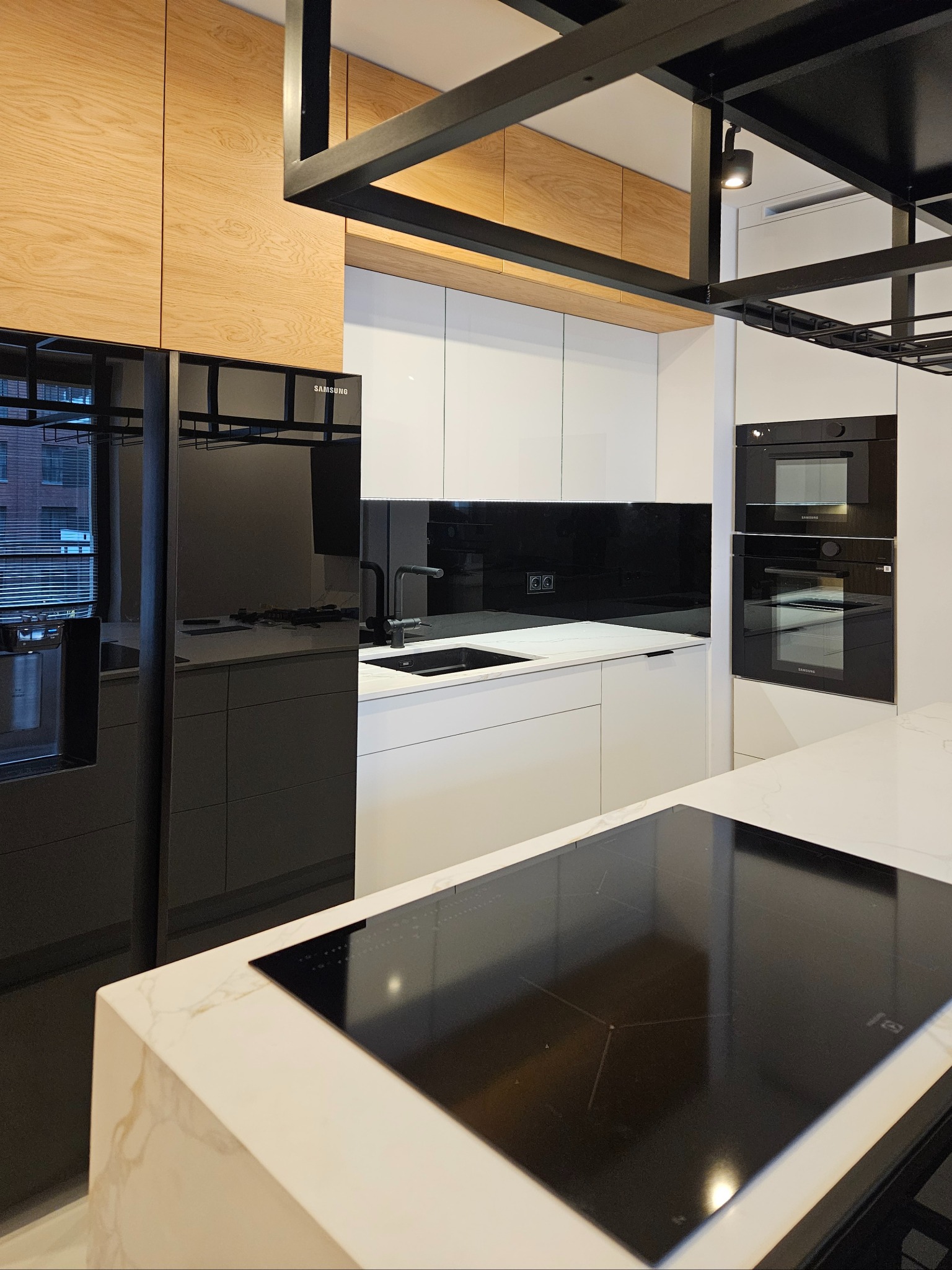 Nowoczesna kuchnia z białymi szafkami, czarnym panelem i sprzętem AGD Samsung. Wyspa z marmurowym blatem i czarną płytą indukcyjną. Drewniane szafki wiszące.