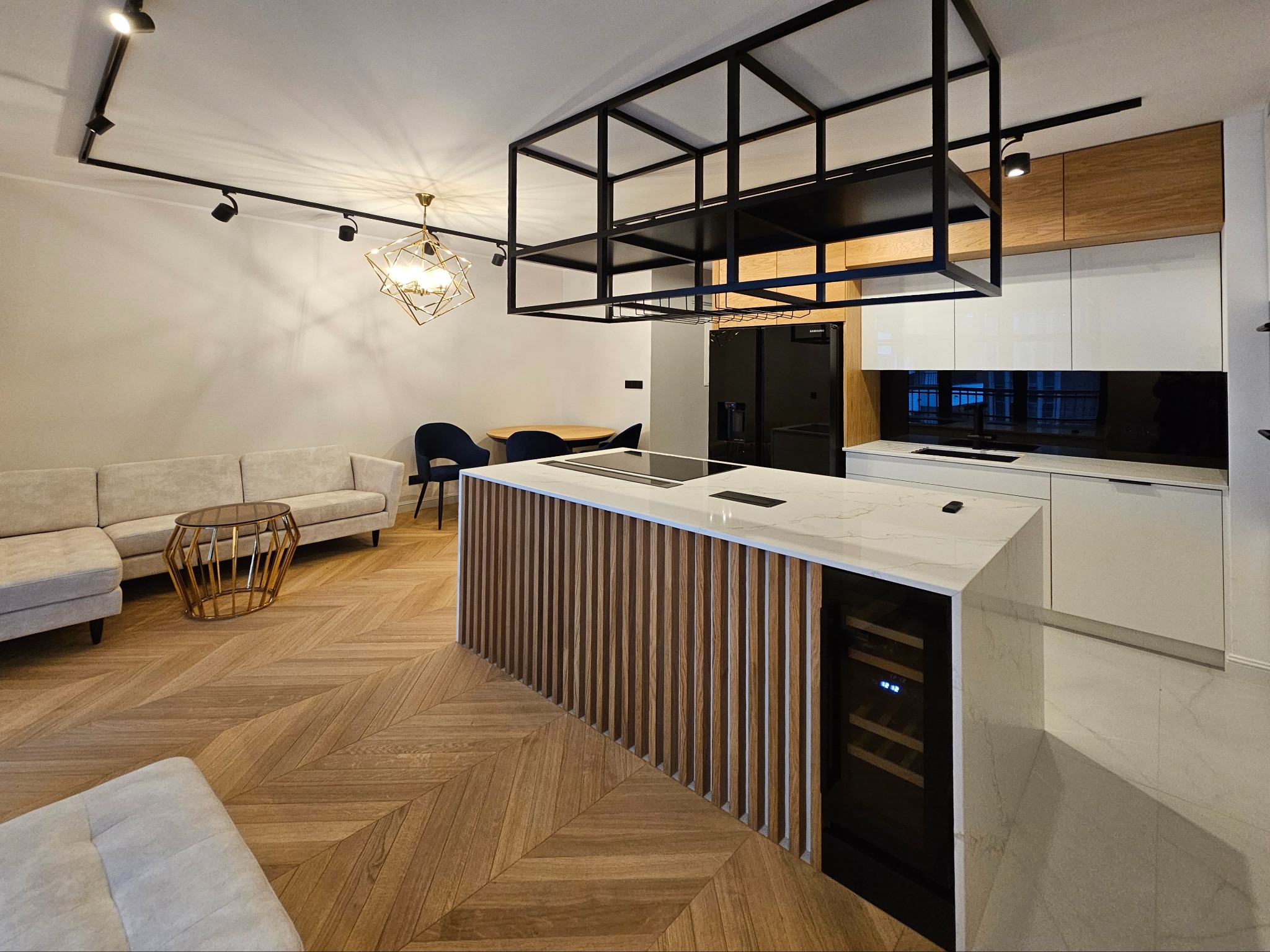 Nowoczesna kuchnia z wyspą o drewnianym froncie i białym blacie. Nad wyspą czarna, geometryczna konstrukcja. Parkiet w jodełkę. Stonowana kolorystyka.