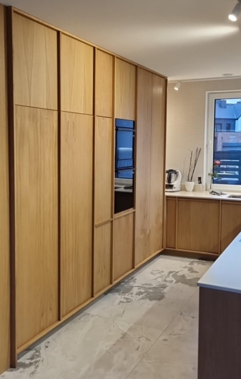 Nowoczesna kuchnia z zabudową z jasnego drewna i wbudowanym piekarnikiem. Minimalistyczny design, marmurowe płytki podłogowe i subtelne oświetlenie punktowe.