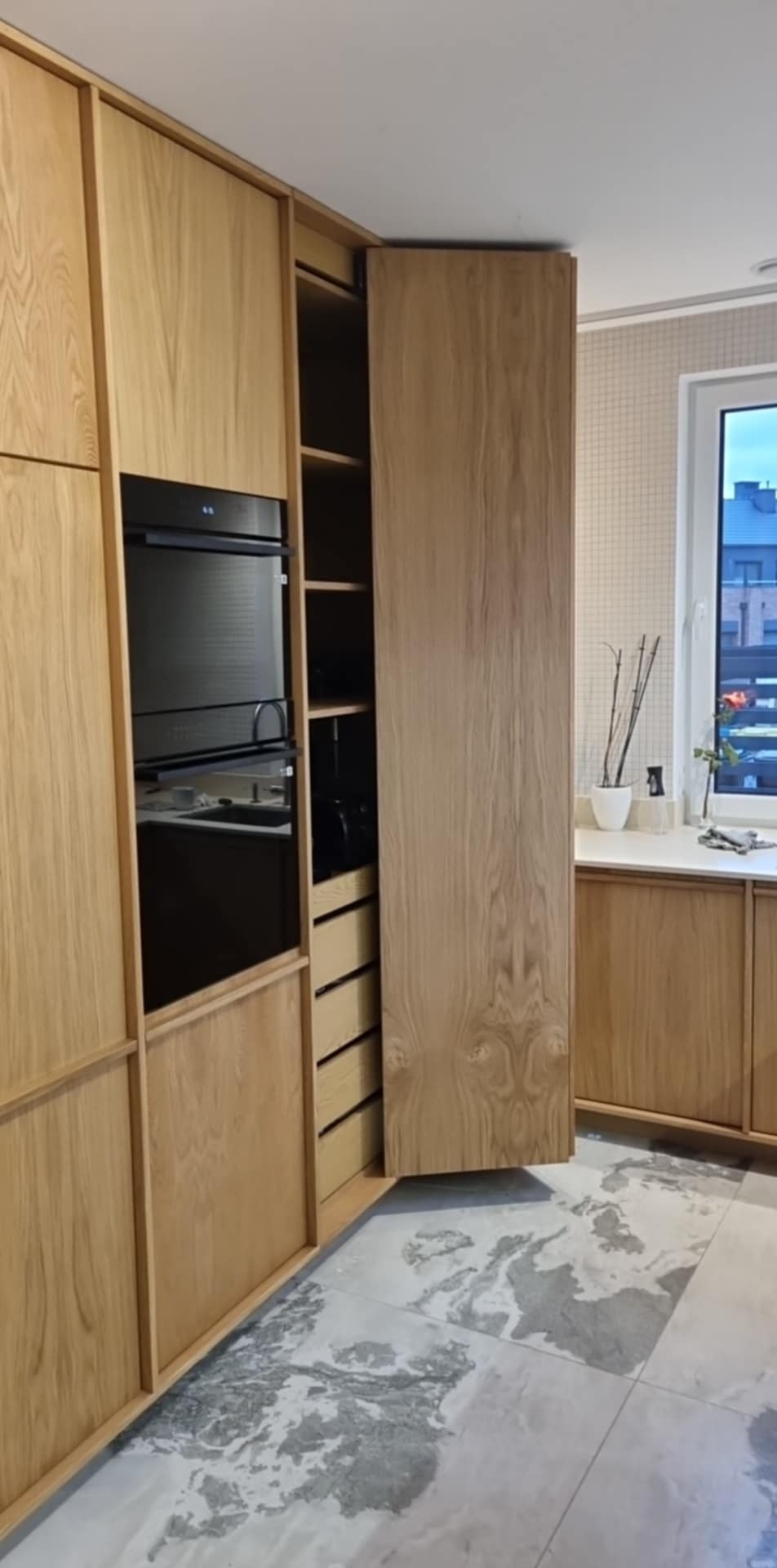 Kuchnia na wymiar z drewna dębowego: zabudowa z piekarnikiem i szafką cargo z wysuwanym frontem. Jasna podłoga z marmurkowym wzorem.
