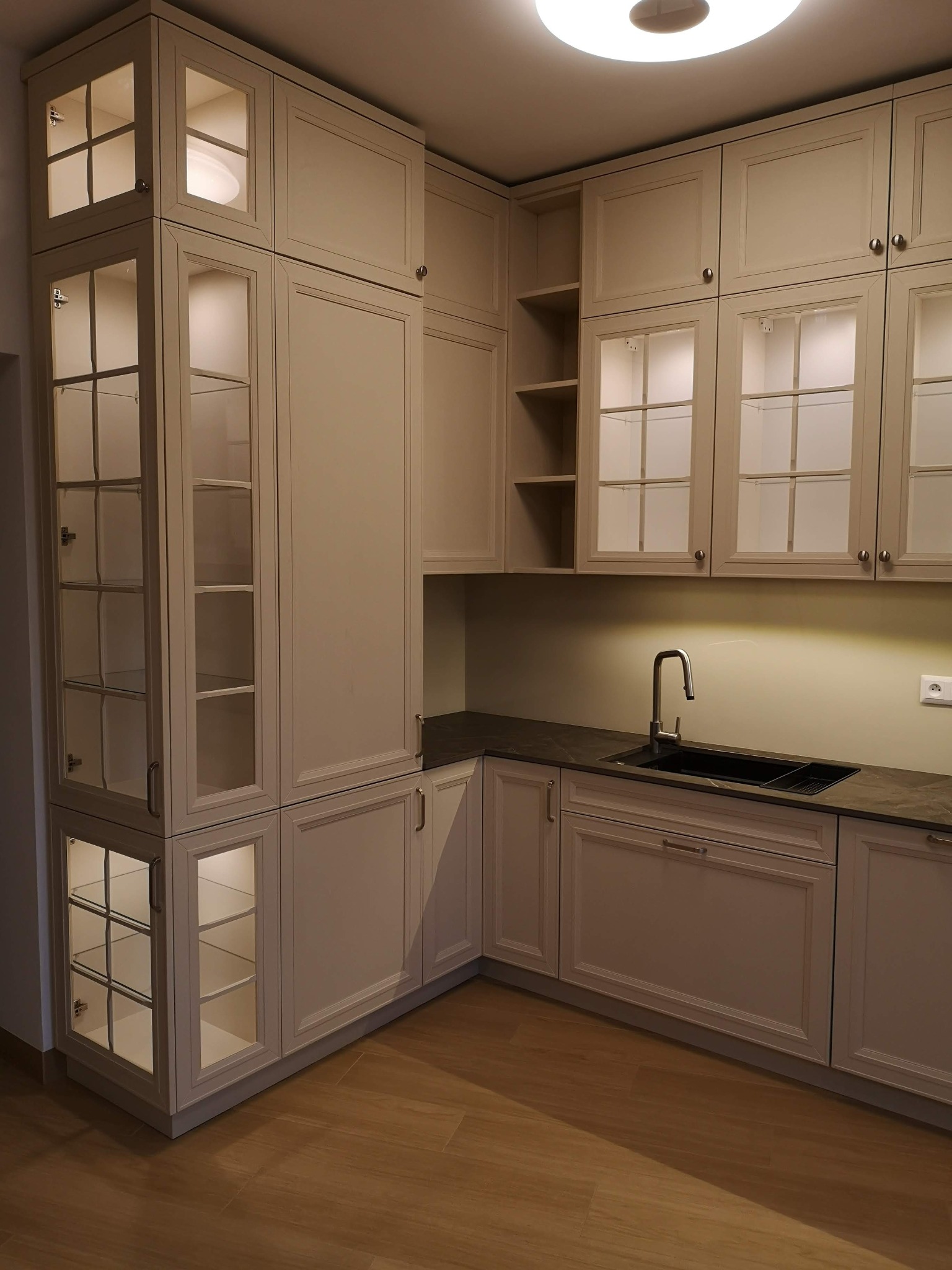 Elegancka, jasna kuchnia z przeszklonymi szafkami i oświetleniem LED, wykończona w klasycznym stylu z ciemnym blatem i drewnianą podłogą.