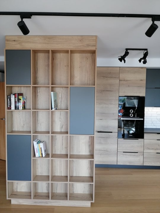 Nowoczesna kuchnia z drewnianą zabudową i regałem z szarymi akcentami, oświetlona reflektorami sufitowymi. Widoczne sprzęty AGD i książki na półkach.