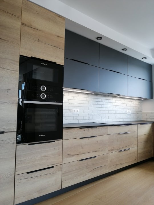 Nowoczesna kuchnia na wymiar: drewnopodobne fronty dolne, grafitowe szafki górne z oświetleniem LED, sprzęt AGD marki Bosch i biała cegła na ścianie.