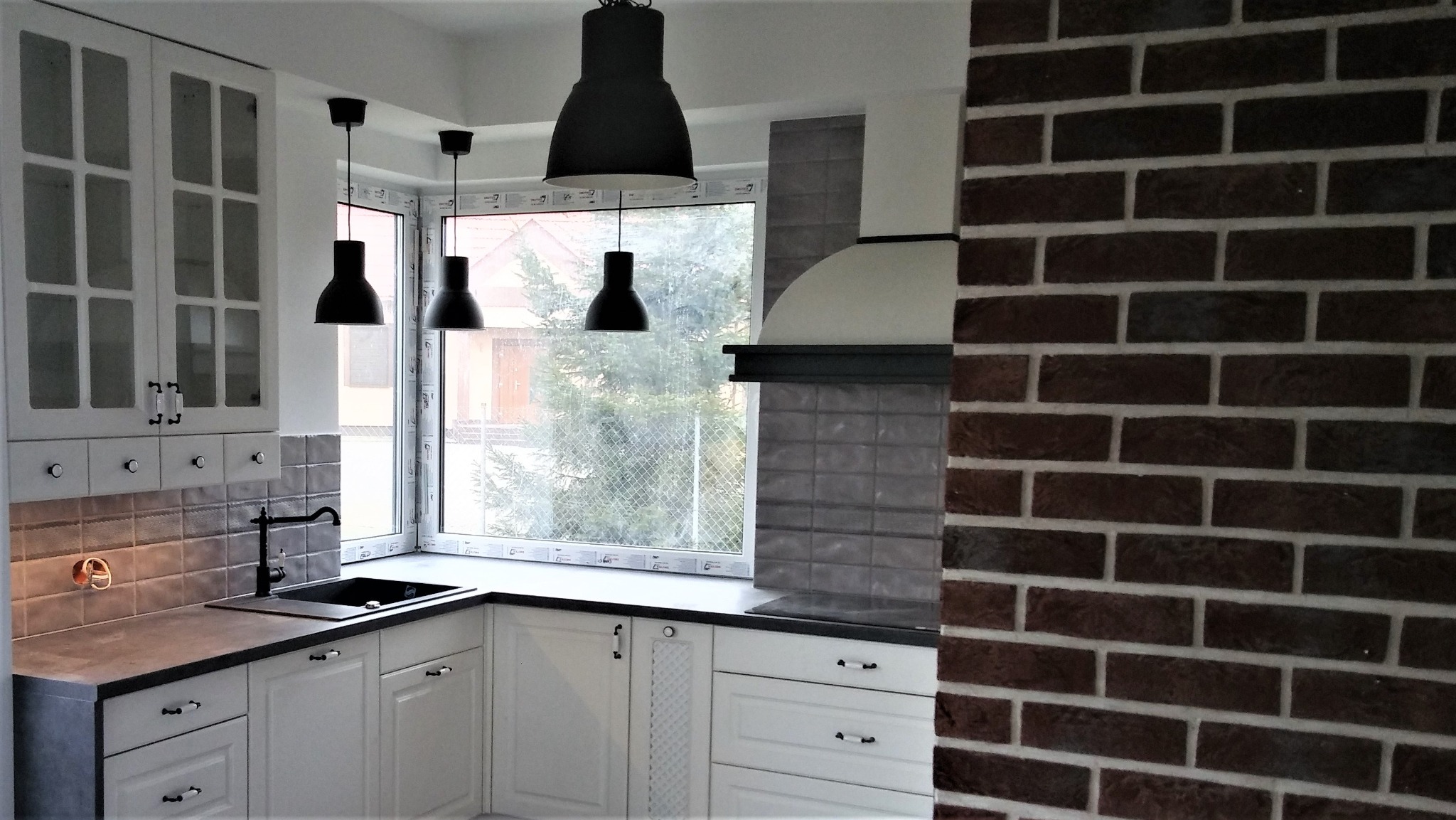 Biała kuchnia z czarnym blatem i lampami, okno narożne z widokiem na zewnątrz, ściana z cegły z prawej strony. Styl rustykalny z nowoczesnymi akcentami.