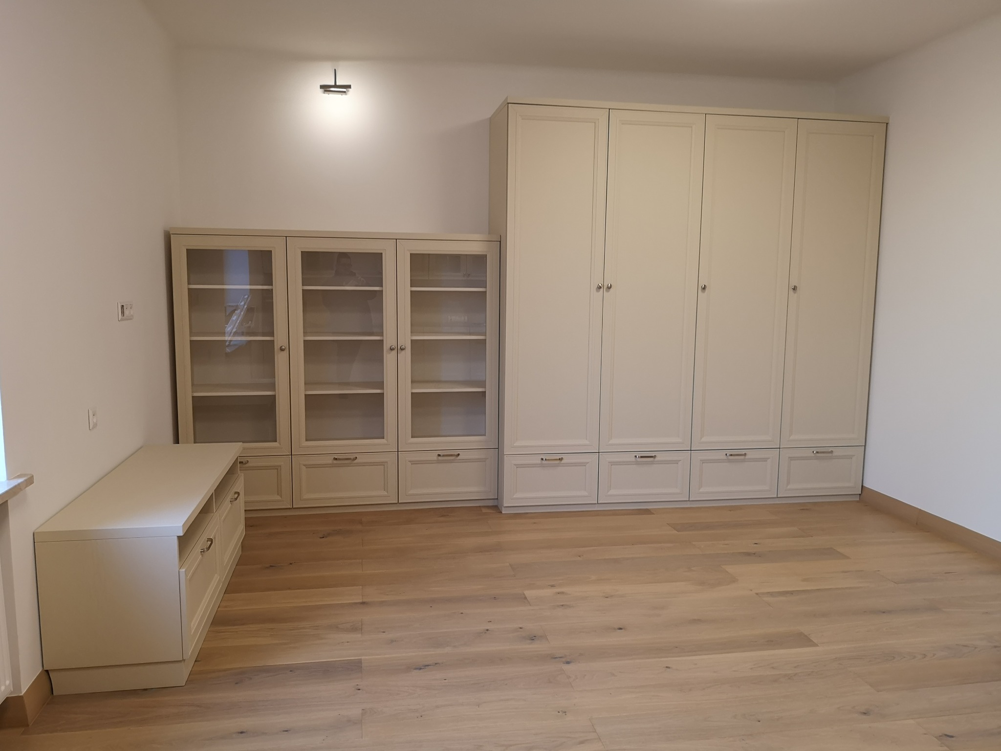 Jasna zabudowa meblowa: szafa, witryna i komoda z szufladami, tworząca spójną aranżację wnętrza na drewnianej podłodze.