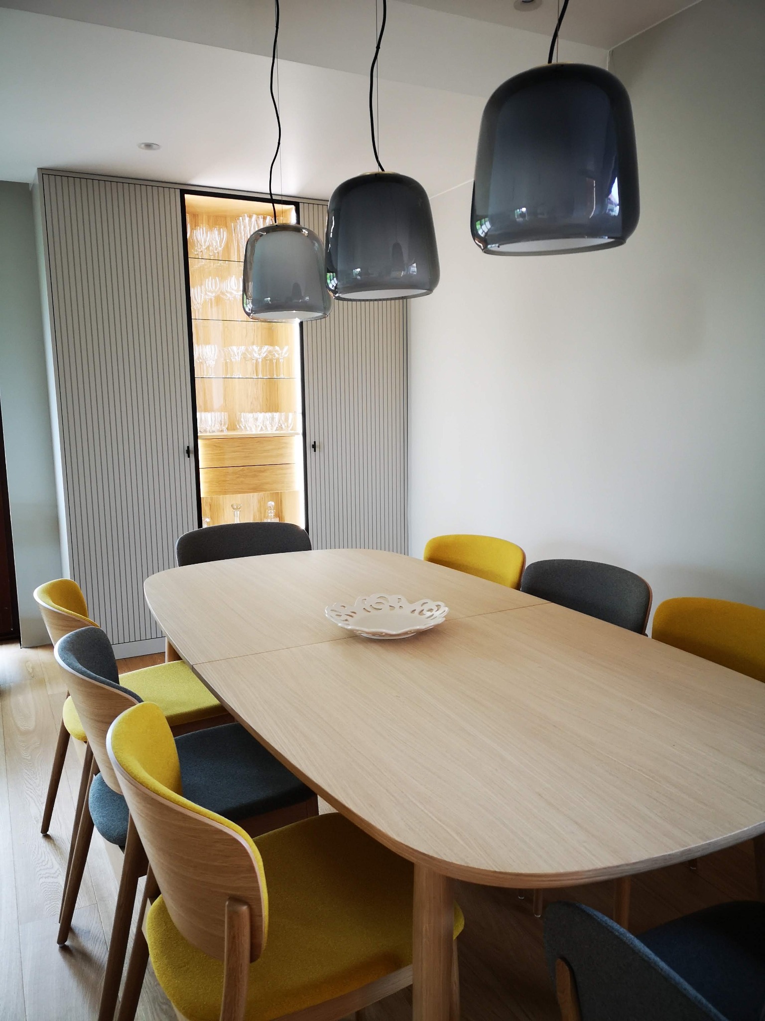 Jadalnia z drewnianym stołem otoczonym krzesłami w odcieniach szarości i żółci. Nad stołem wiszą trzy szare lampy. W tle szafa wnękowa z podświetlonymi szklanymi półkami.