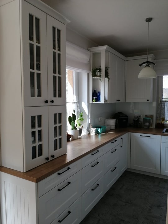 Biała, klasyczna kuchnia z drewnianym blatem i czarnymi uchwytami w Kowalach. Widoczne przeszklone szafki wiszące i rośliny doniczkowe przy oknie.