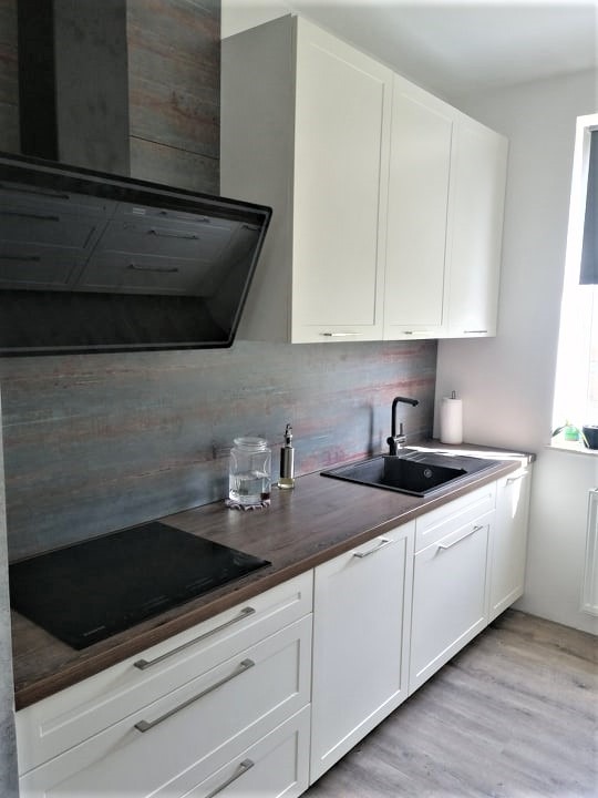 Biała zabudowa kuchenna z drewnianym blatem i czarnym zlewem, nowoczesny okap, szklany dzbanek i drewnopodobne panele podłogowe. Ujęcie z perspektywy.