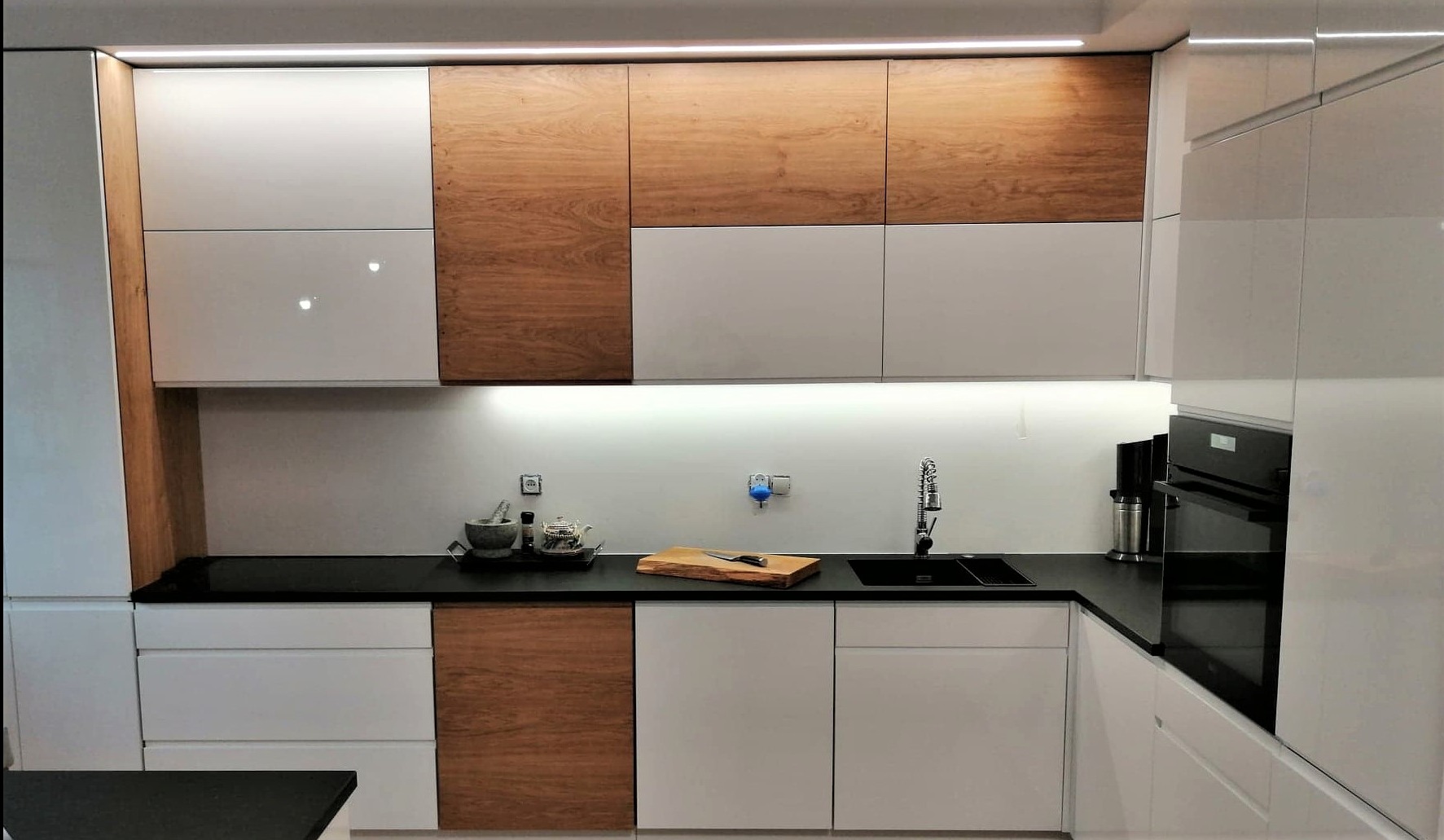 Nowoczesna kuchnia z białymi, lakierowanymi frontami i drewnianymi akcentami. Czarny blat, oświetlenie LED pod szafkami, sprzęt AGD w zabudowie.
