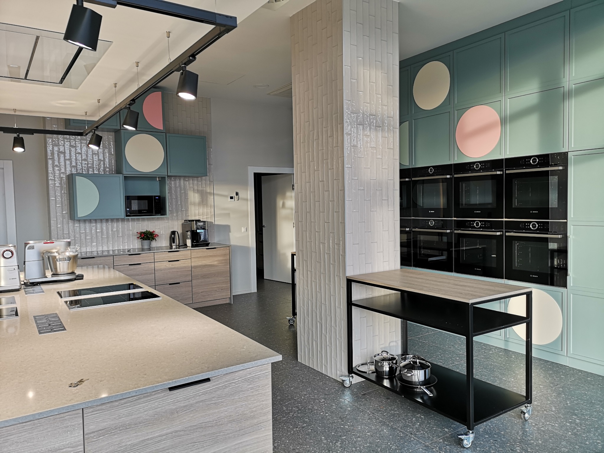 Nowoczesna kuchnia: jasne drewno, pastelowe szafki z okrągłymi akcentami, rząd piekarników Bosch. Minimalistyczny design, oświetlenie szynowe, wyspa kuchenna z płytą indukcyjną.