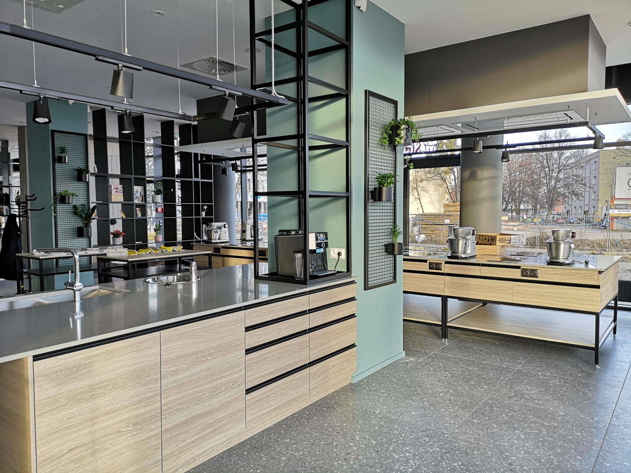 Nowoczesna kuchnia z wyspą i elementami metalowymi. Jasne drewno frontów szafek kontrastuje z szarym blatem i podłogą. Widoczne oświetlenie szynowe i dekoracje roślinne.