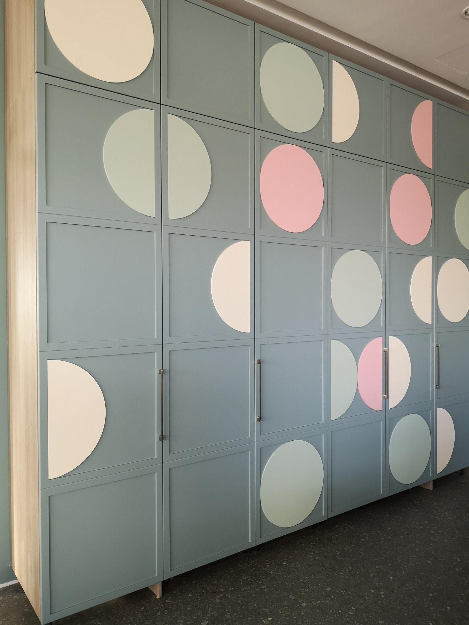 Niebieska szafa z geometrycznym wzorem z kolorowych kół, widoczna z perspektywy. Minimalistyczny design, metalowe uchwyty, jasne tło.