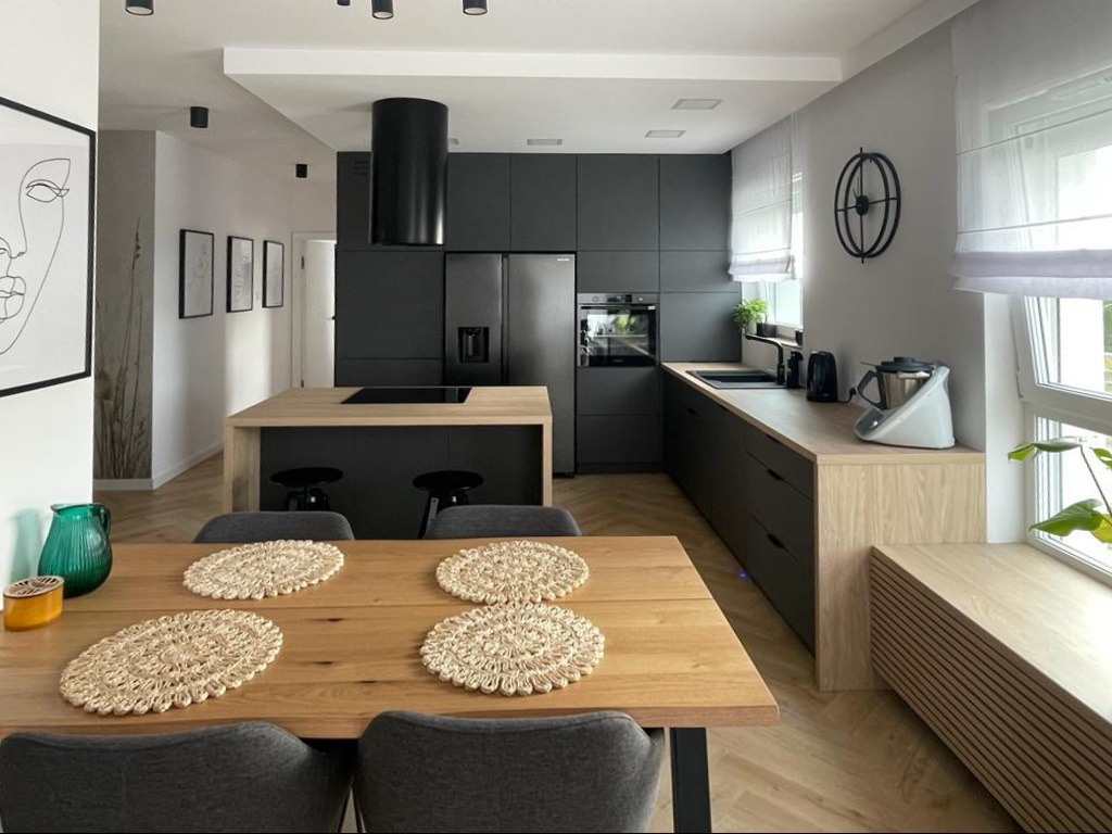 Nowoczesna kuchnia w odcieniach szarości i drewna: zabudowa kuchenna z wyspą, minimalistyczne dodatki, drewniany stół z podkładkami. Jasne wnętrze z dużym oknem.