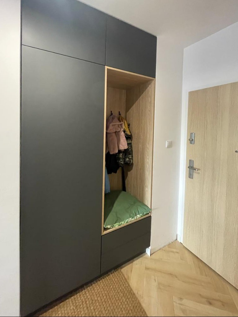 Nowoczesna szafa wnękowa z siedziskiem i wieszakami, w odcieniach szarości i drewna, w przedpokoju z parkietem w jodełkę. Minimalistyczny design.
