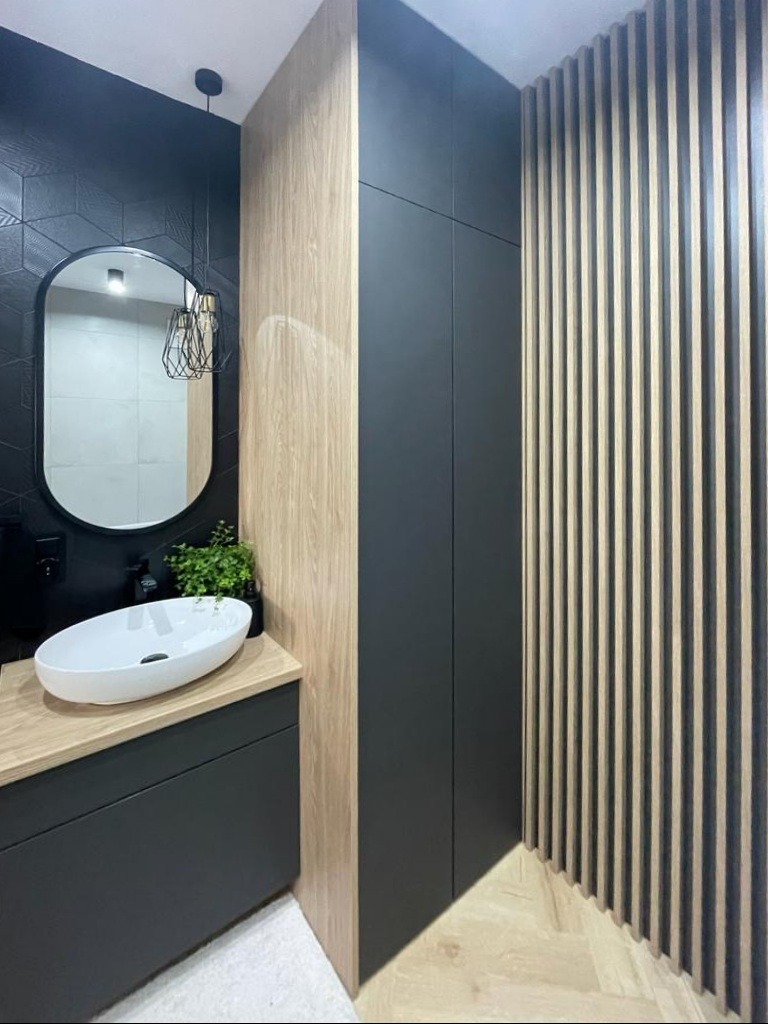 Nowoczesna łazienka z owalnym lustrem, umywalką nablatową i drewnianymi lamelami. Ciemne płytki i szafki tworzą elegancki kontrast z jasnym drewnem.