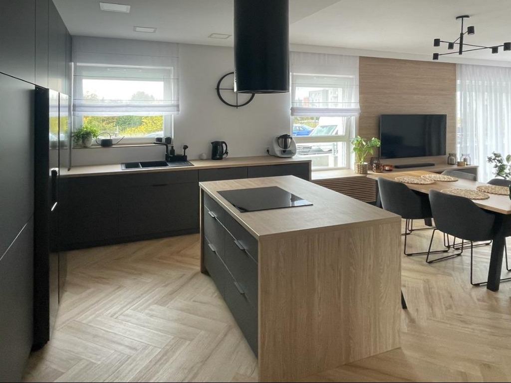 Nowoczesna kuchnia z wyspą i czarnymi szafkami, jasny parkiet w jodełkę. Okno z roletami rzymskimi, minimalistyczny okap, stół z krzesłami.