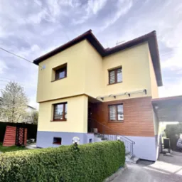 Dwukondygnacyjny dom z żółtą elewacją, brązowymi oknami i drewnianą okładziną na dole, sfotografowany w pochmurny dzień z widocznym fragmentem ogrodu i żywopłotu z lampami.