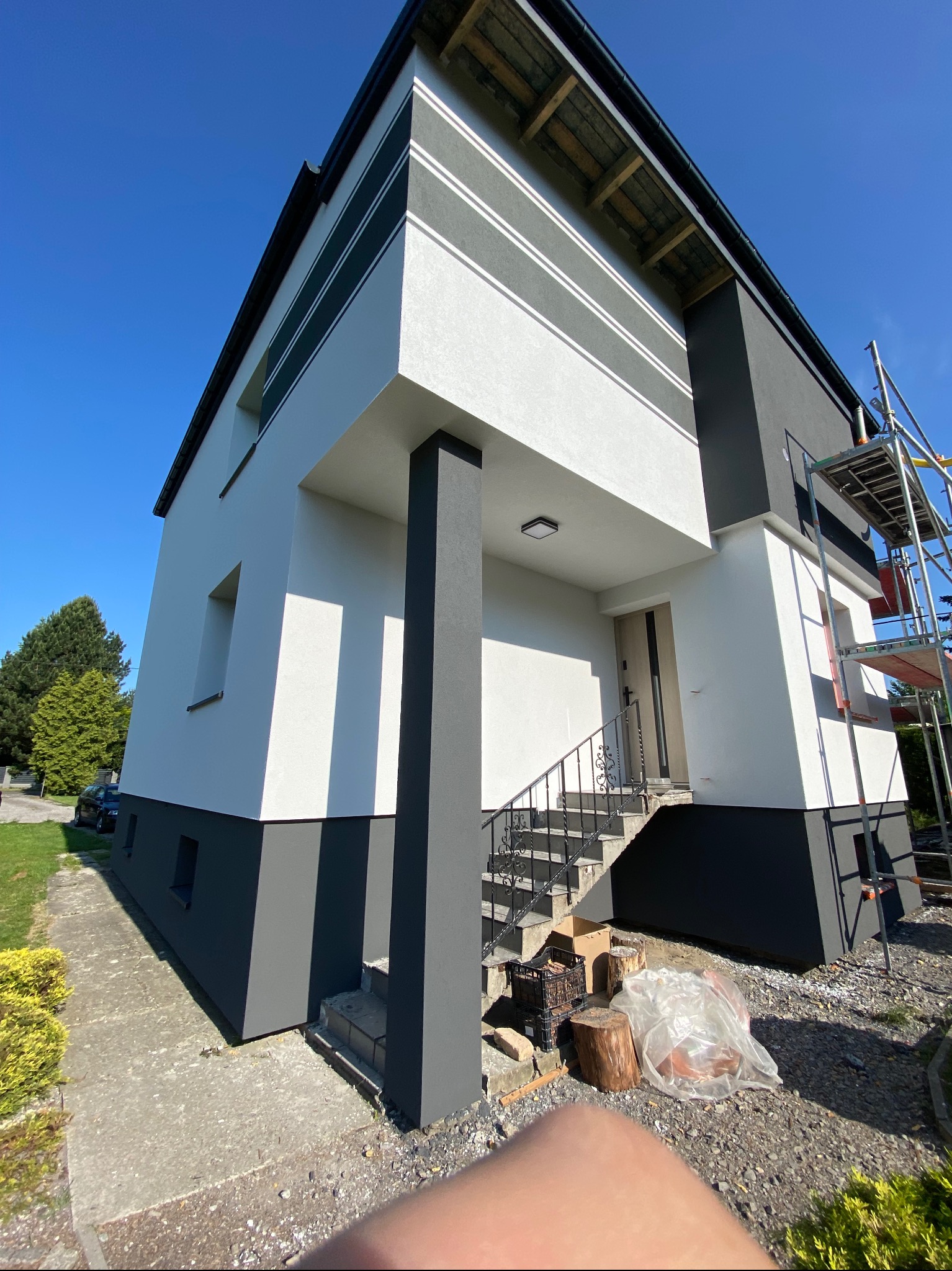 Dwukondygnacyjny dom jednorodzinny z nową, biało-szarą elewacją, widoczny z dołu, z elementami dekoracyjnymi w postaci poziomych pasów. Częściowo widoczne rusztowanie. Na pierwszym planie fragment...