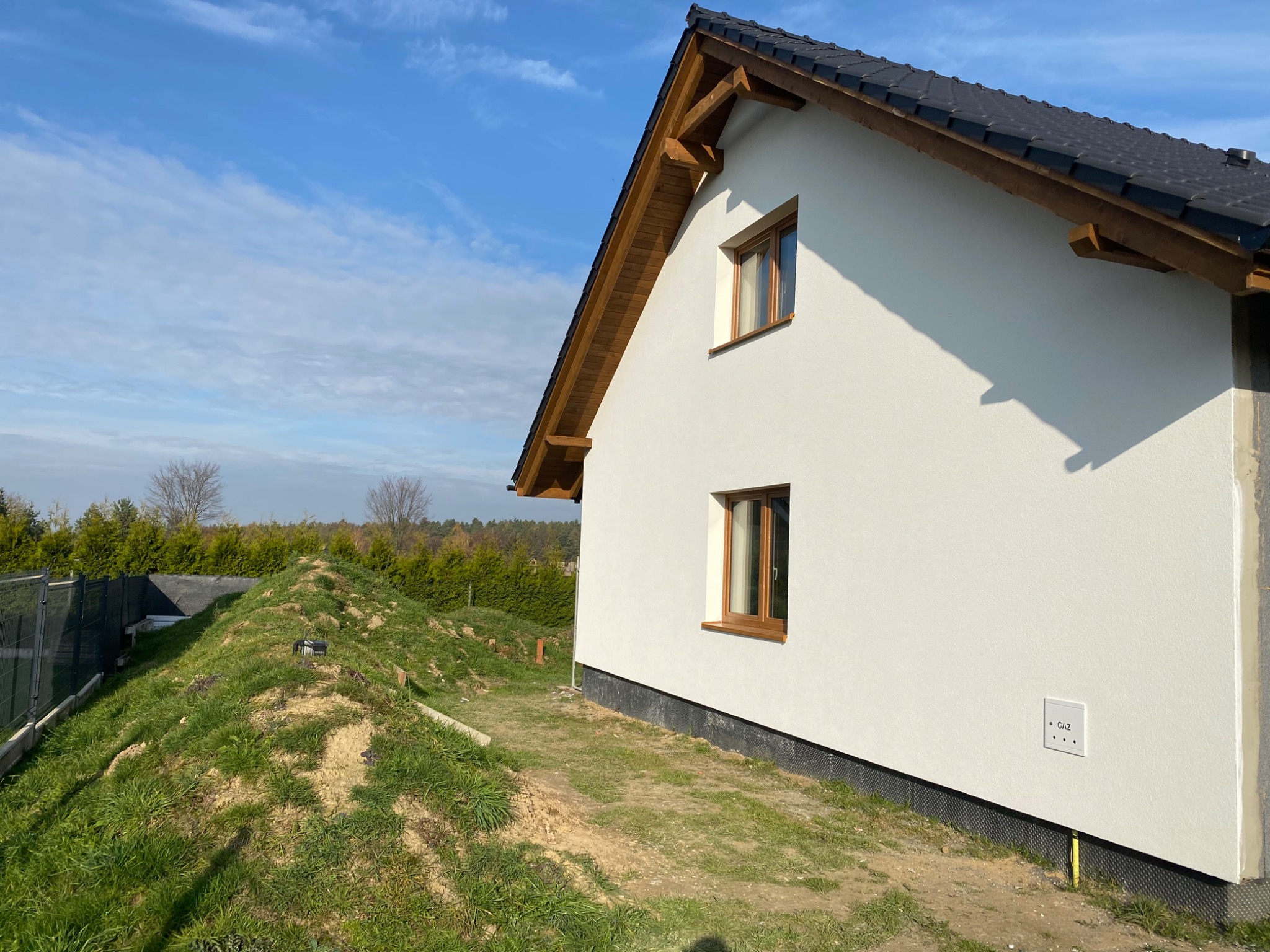 Boczny widok domu z białą elewacją i drewnianymi elementami wykończeniowymi dachu oraz okien, na tle błękitnego nieba i zieleni, widoczna skrzynka gazowa na elewacji.