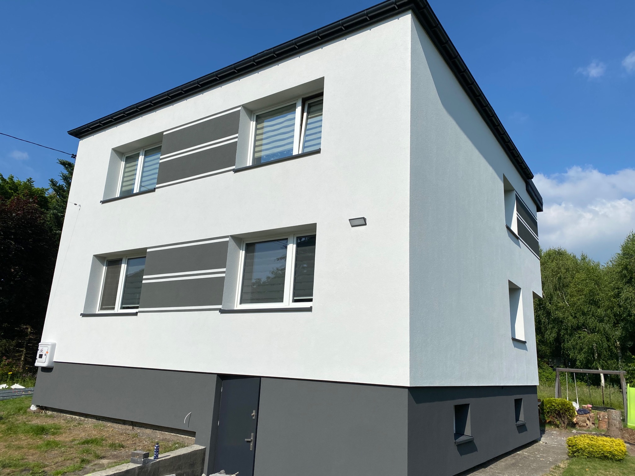 Dwukondygnacyjny budynek z nowoczesną elewacją w odcieniach bieli i szarości, z widocznymi poziomymi pasami dekoracyjnymi wokół okien, sfotografowany w słoneczny dzień.