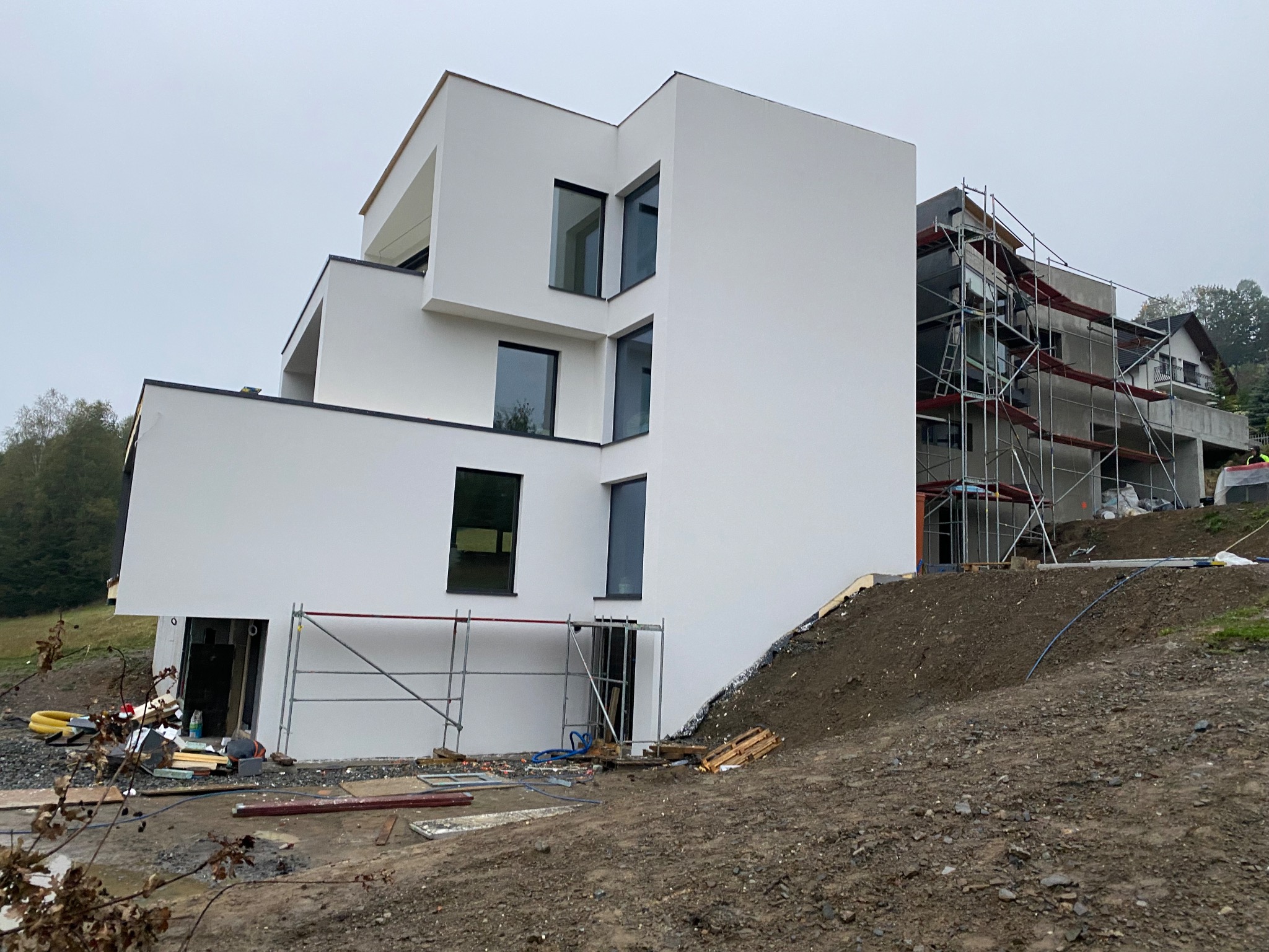 Nowoczesny, trzypiętrowy dom z białą elewacją w trakcie prac wykończeniowych, widoczne rusztowania i przygotowany teren wokół budynku.