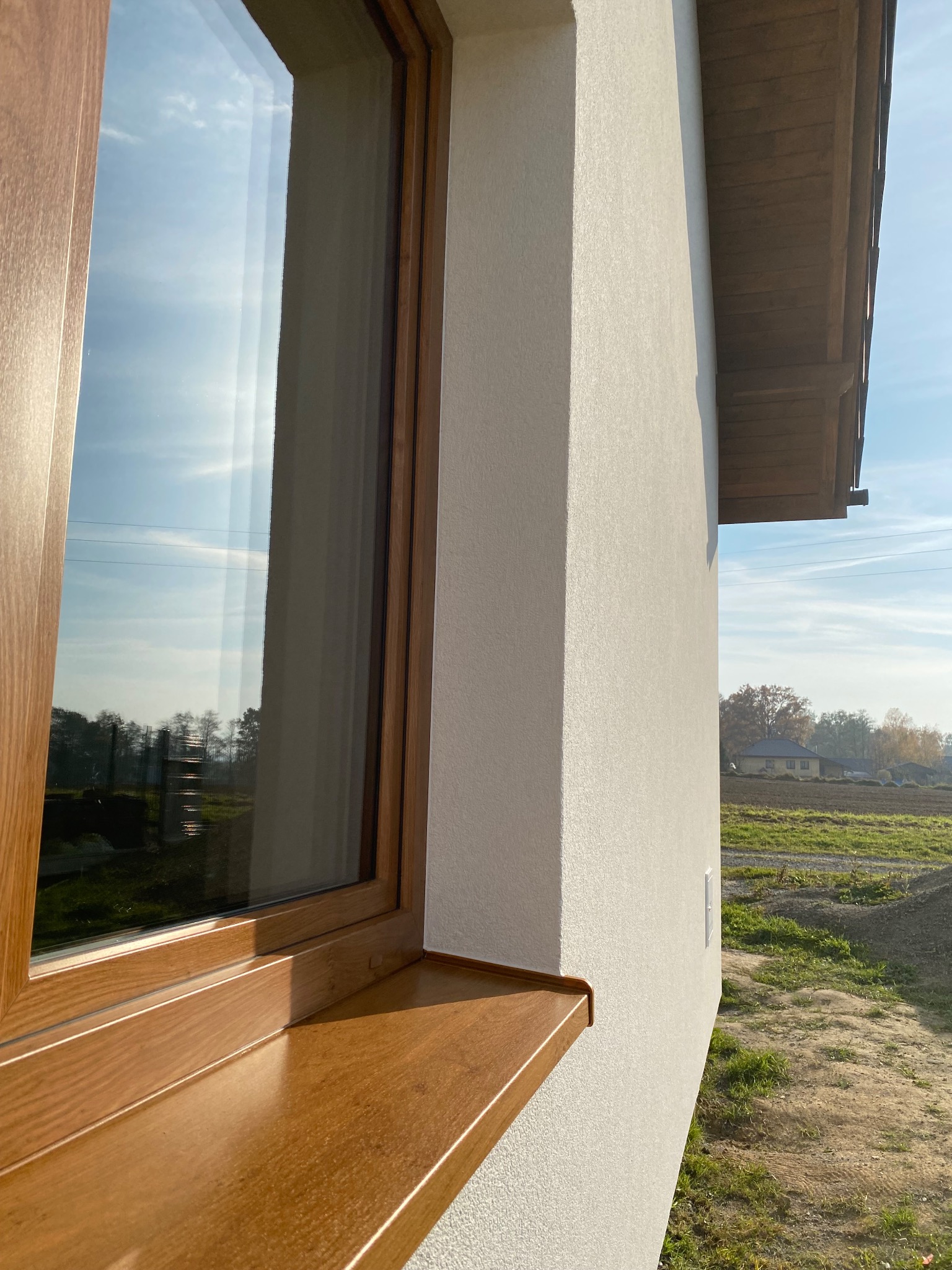 Fragment elewacji domu po nałożeniu tynku strukturalnego, widoczne okno z drewnopodobną ramą i parapetem oraz fragment dachu.