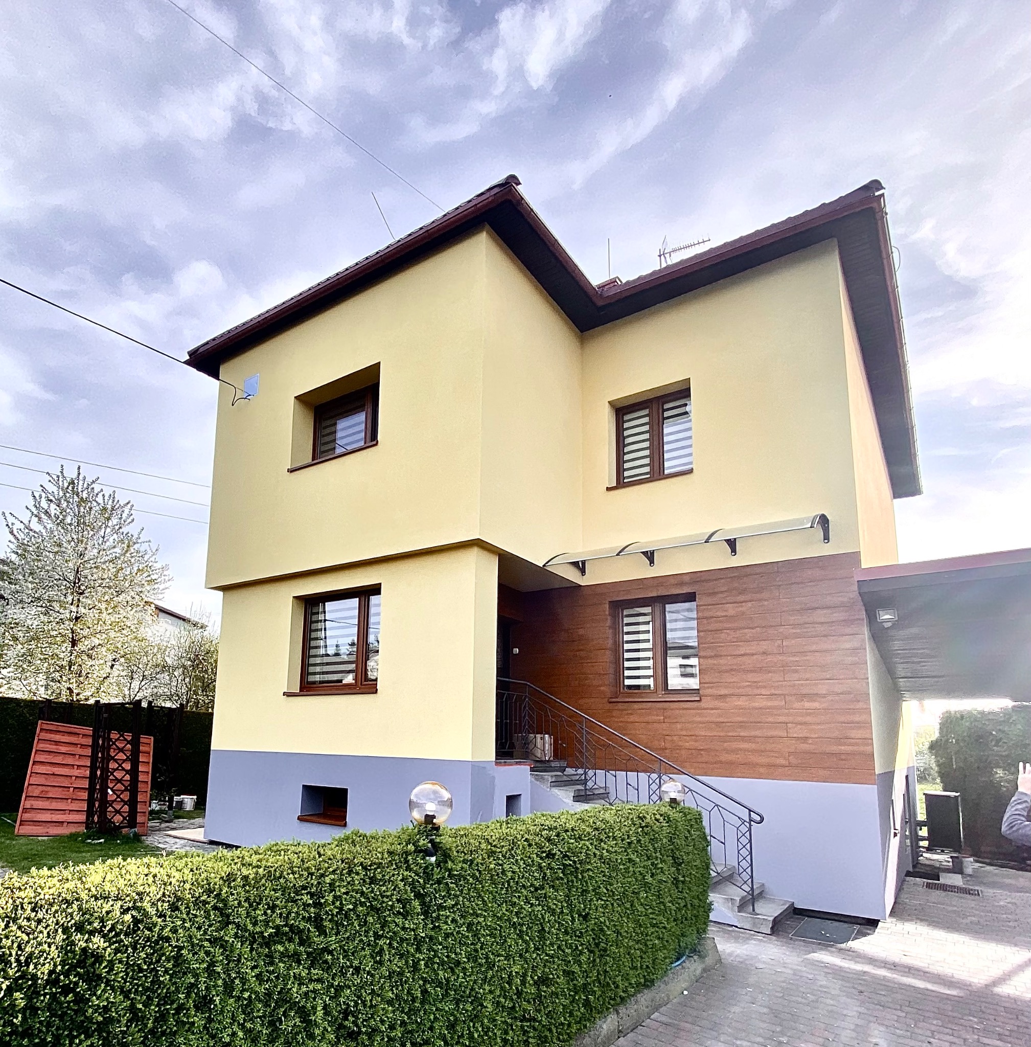 Dwukondygnacyjny dom z żółtą elewacją, brązowymi oknami i drewnianą okładziną na dole, sfotografowany w pochmurny dzień z widocznym fragmentem ogrodu i żywopłotu z lampami.