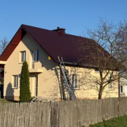 Dwupiętrowy dom z brązowym dachem widziany zza drewnianego płotu. Do dachu przystawiona aluminiowa drabina.