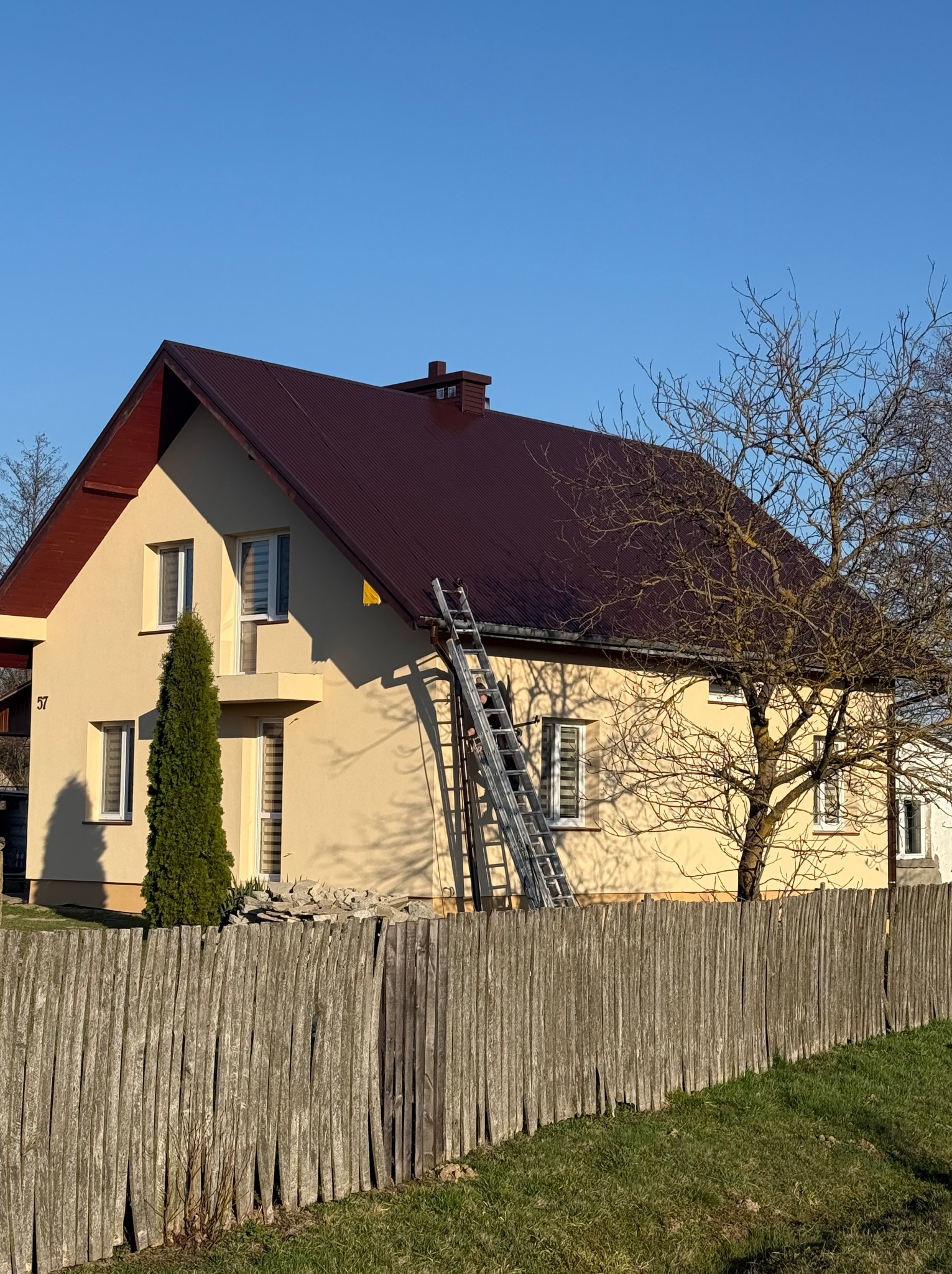 Dwupiętrowy dom z brązowym dachem widziany zza drewnianego płotu. Do dachu przystawiona aluminiowa drabina.