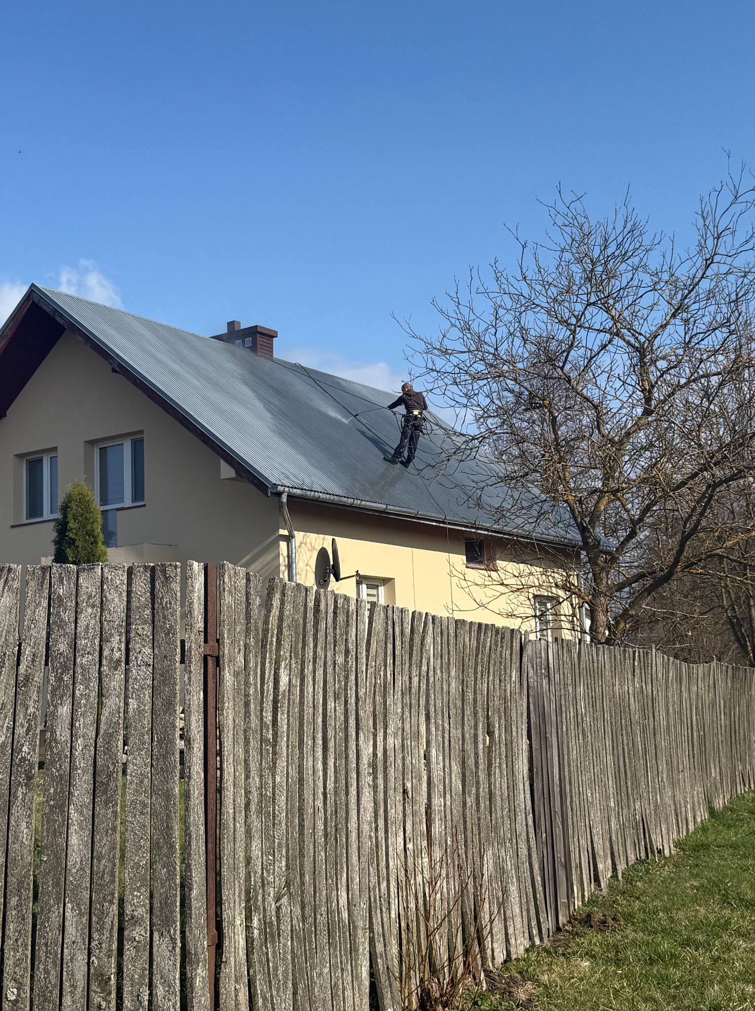 Pracownik w uprzęży na dachu domu jednorodzinnego, wykonuje prace na wysokości, widok zza drewnianego płotu.