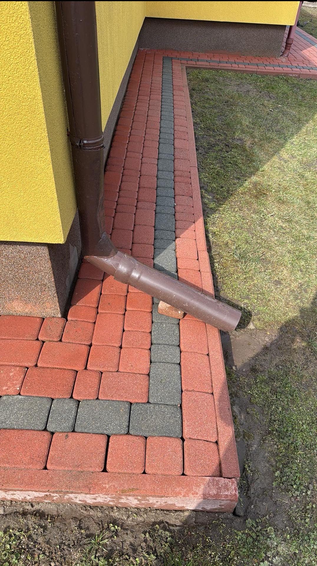 Fragment elewacji budynku z żółtą ścianą i brązowym orynnowaniem, obok ułożona kostka brukowa w kolorach czerwonym i szarym, z fragmentem rynny leżącej na kostce.