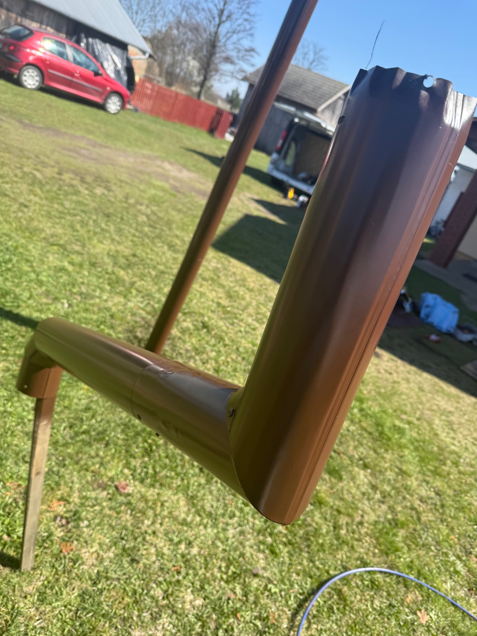 Brązowa rynna dachowa z tworzywa sztucznego, tymczasowo podparta drewnianymi kołkami na trawniku w słoneczny dzień, z widocznym fragmentem niebieskiego węża ogrodowego w tle.