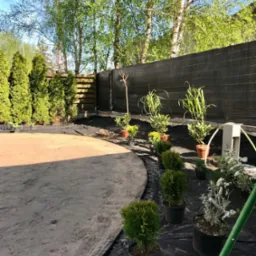 Przygotowany teren pod przyszły ogród z rozłożoną agrowłókniną, obrzeżami i roślinami w donicach wzdłuż ogrodzenia.