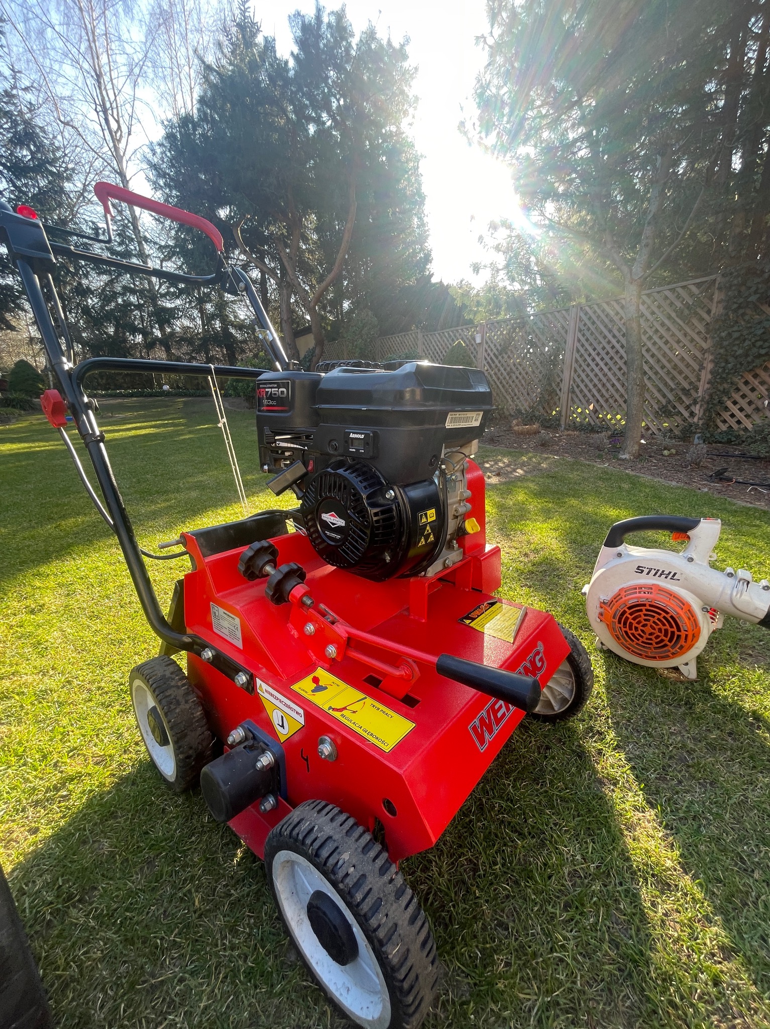 Czerwony aerator spalinowy WEMG na trawniku, obok dmuchawa Stihl, w tle drzewa i płot, oświetlone słońcem.