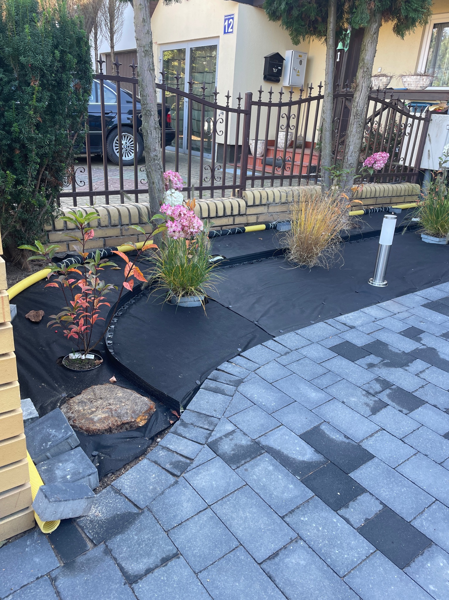 Fragment ogrodu w trakcie tworzenia: widoczne sadzonki w doniczkach, czarna agrowłóknina, obrzeża, kostka brukowa, lampa ogrodowa i elementy instalacji nawadniającej.